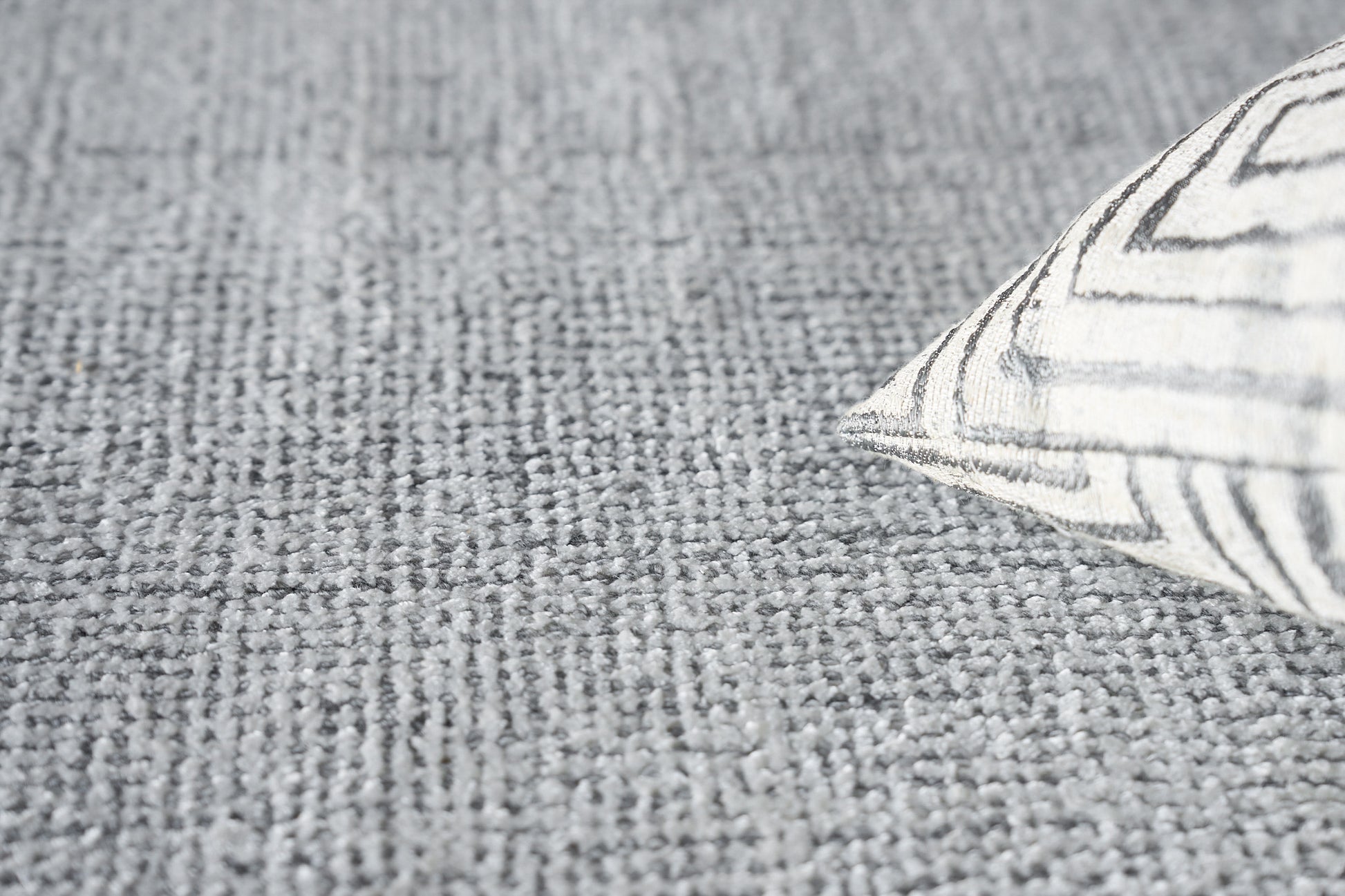 Close-up of a grey textured woven rug with a white cushion, showing a bold grey geometric pattern.