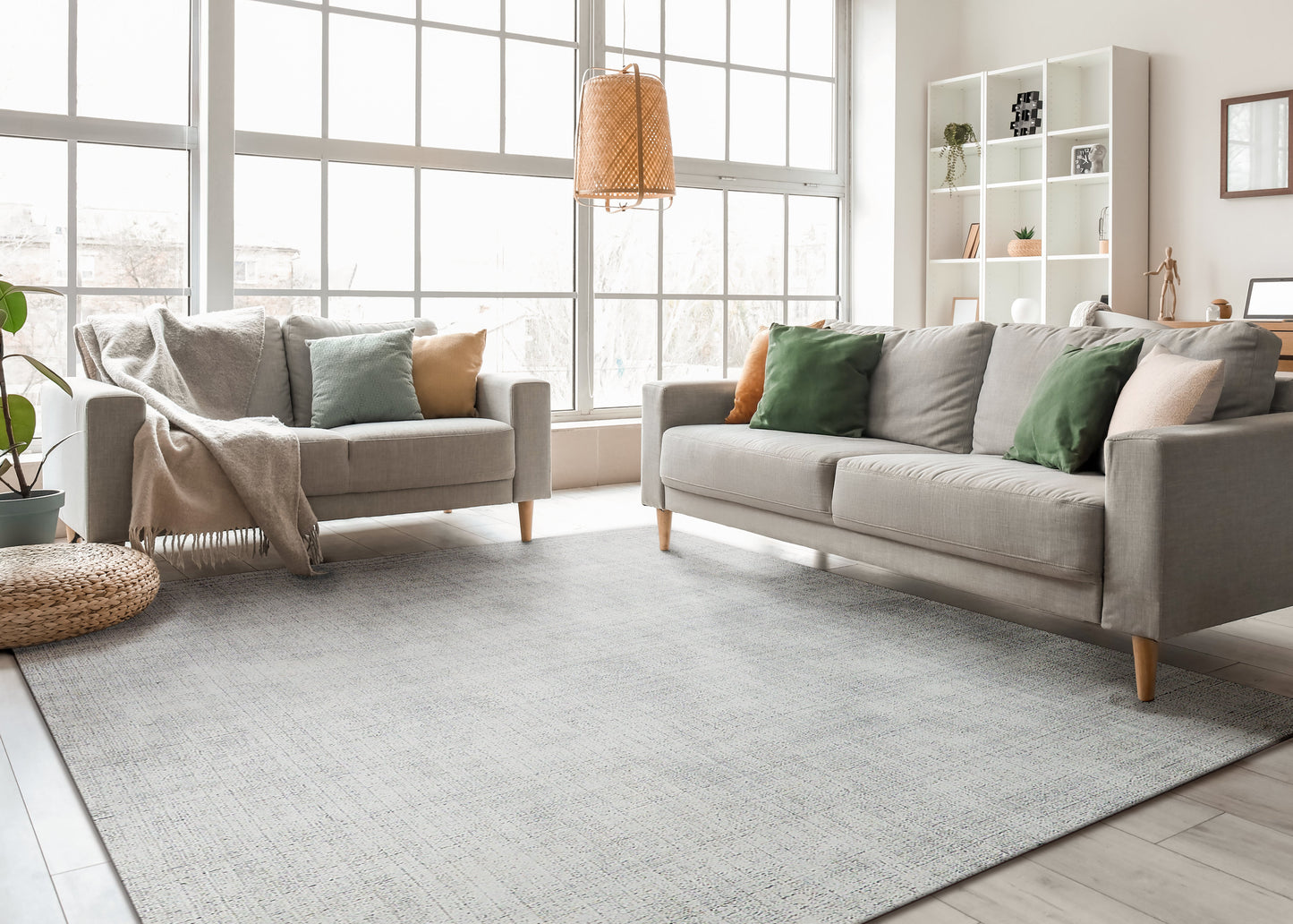 Bright living room with two light gray sofas, green/orange cushions, large windows, and a soft gray area rug.
