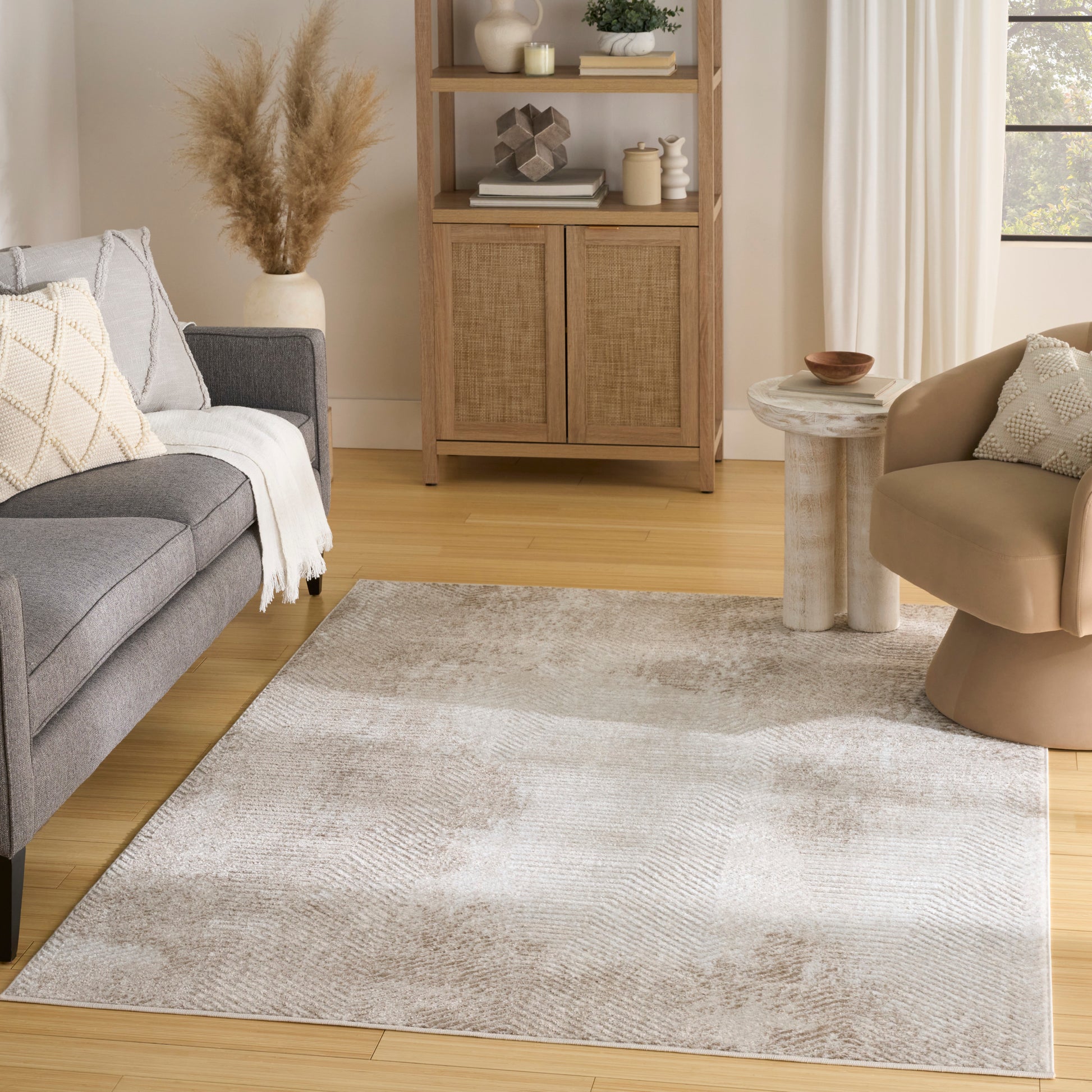 Neutral living room with light beige rug, gray sofa, tan curved armchair, wooden cabinet, pampas plant, and round side table.