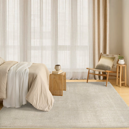Neutral bedroom with beige linen bedding, large window, sheer curtains, light gray rug, and wooden slat side table with woven rattan armchair.