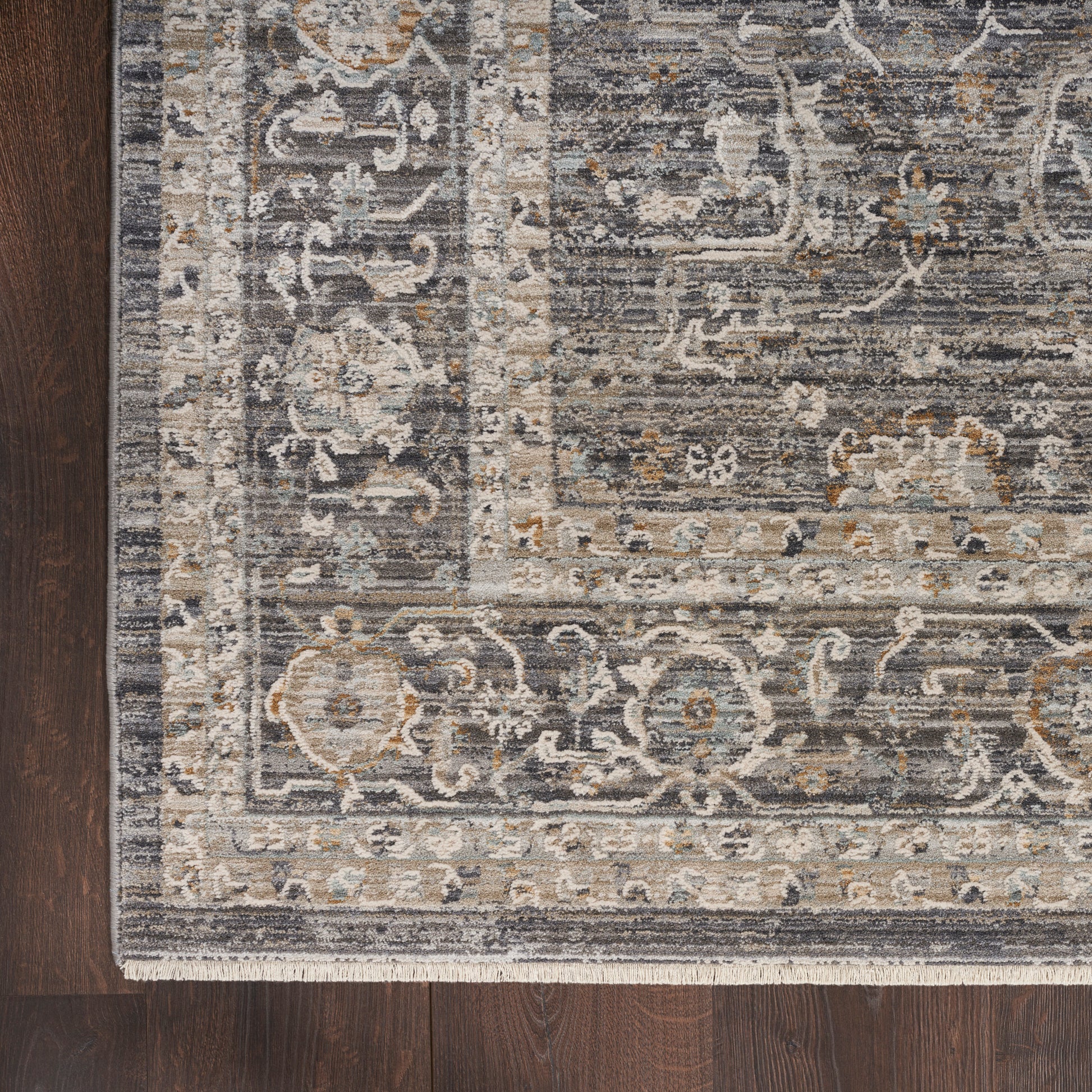 Vintage-style grey and ivory patterned area rug with blue and tan accents and fringed edge on dark hardwood floor.