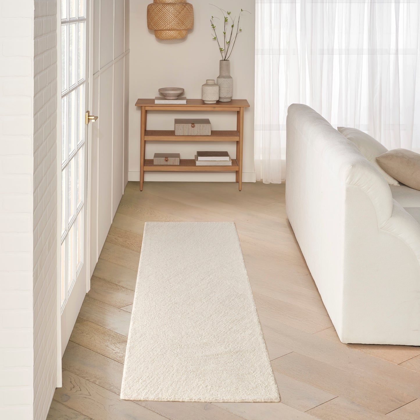 Cream hallway runner rug, pale wood floor, white sofa on the right, & light wood console with baskets & vases.