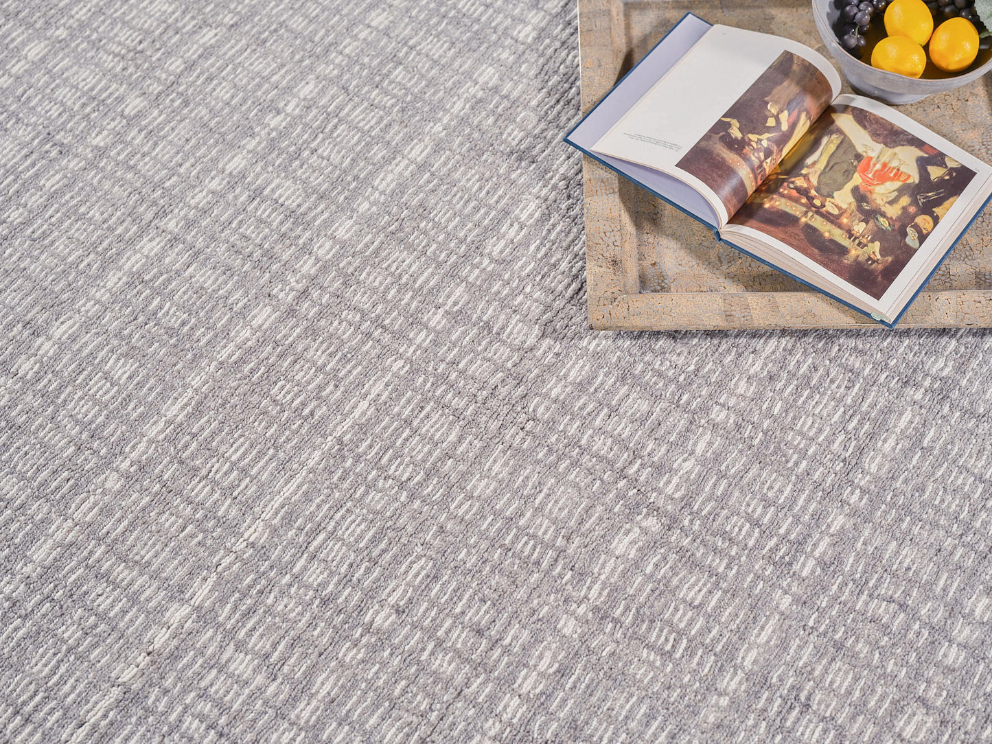 Grey textured rug with a subtle linear pattern; wooden tray with an open art book and a bowl of lemons and grapes.