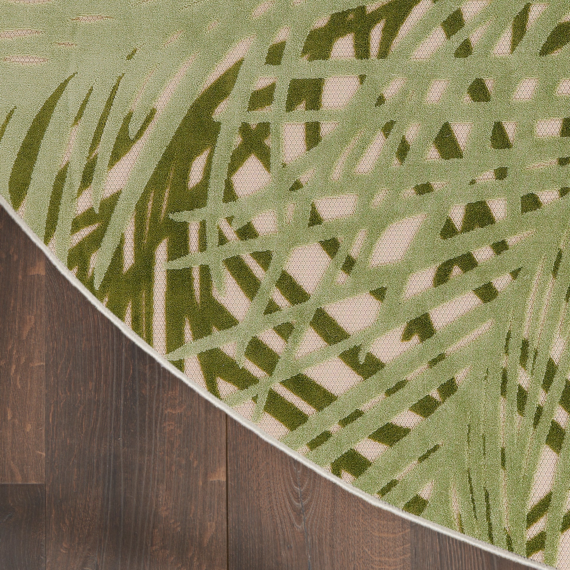 Green abstract-pattern round outdoor rug with beige field, edge curling over dark wood floor.