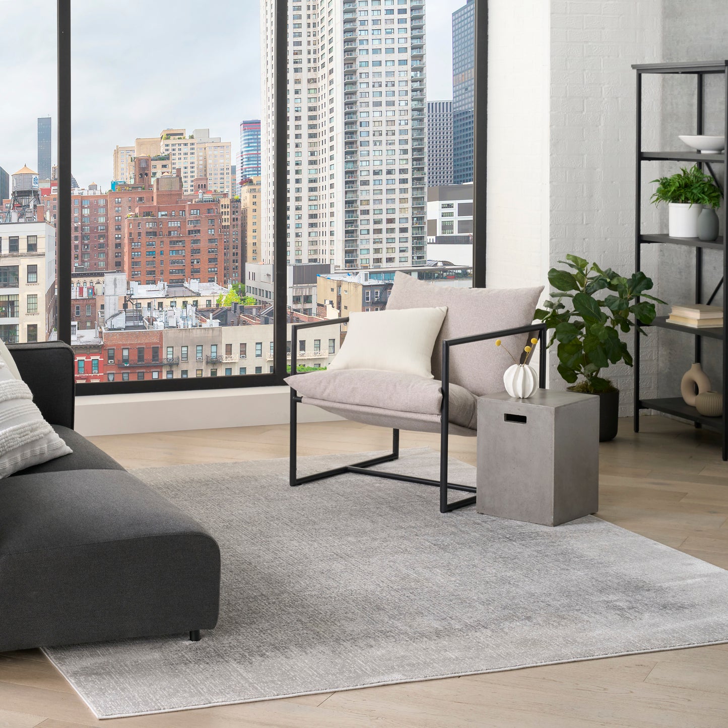 Contemporary living room with city view, gray sofa, light rug, metal-framed armchair, concrete side table and plants.