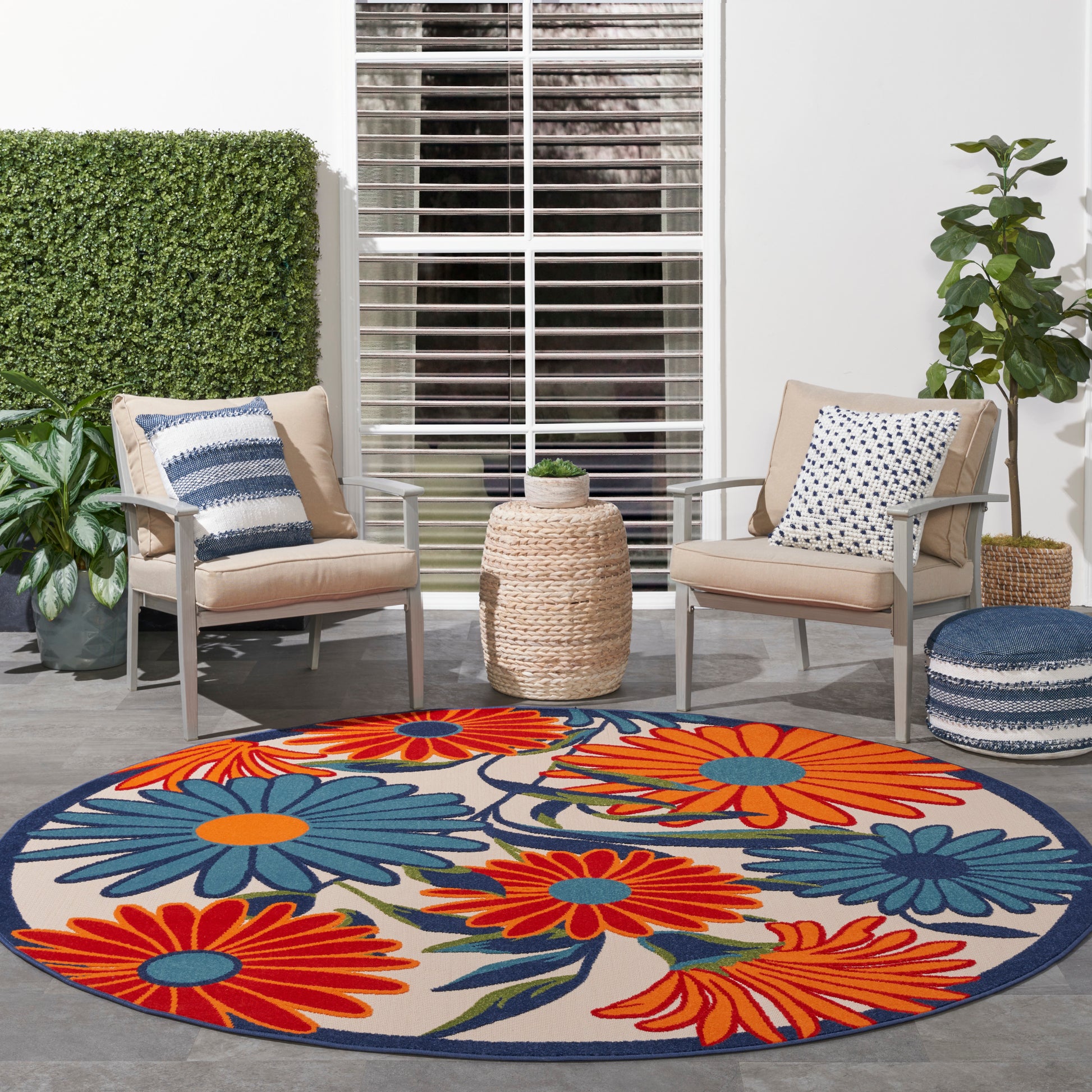 Round outdoor rug with bold orange, blue & red flowers and navy border; beige patio chairs, round wicker pouf, potted plants.
