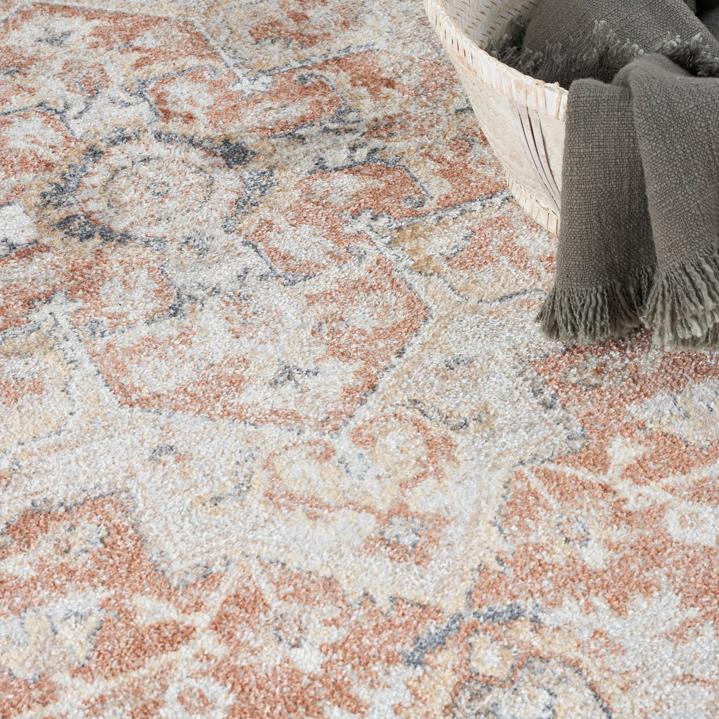 Muted peach, beige and gray patterned area rug with a woven basket and a dark gray fringed throw blanket.