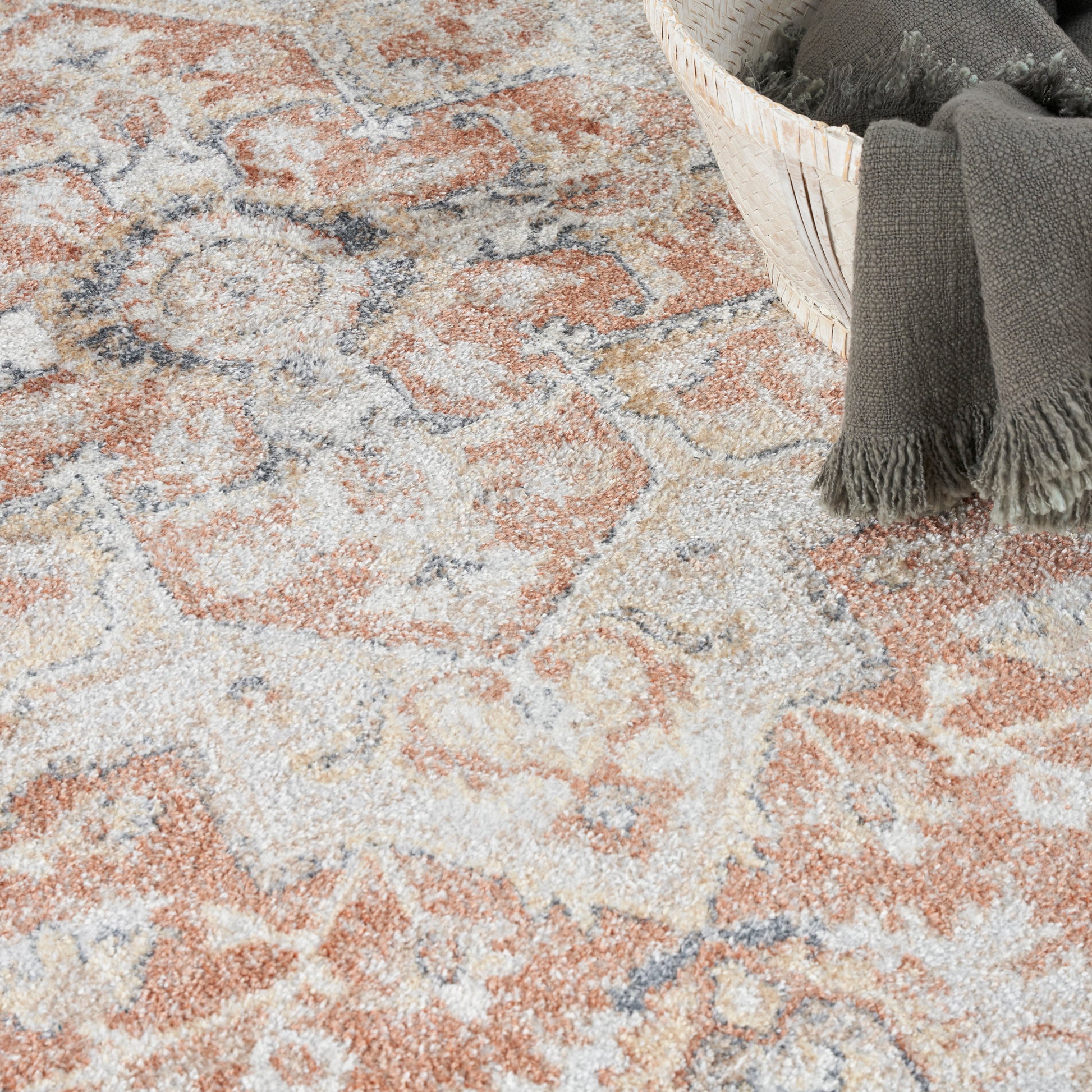 Muted peach, beige and gray patterned area rug with a woven basket and a dark gray fringed throw blanket.