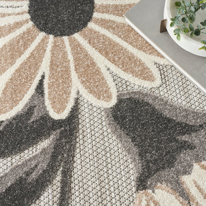 Neutral floral rug with large beige petals and charcoal center on a textured gray weave; plant and tray top-right.
