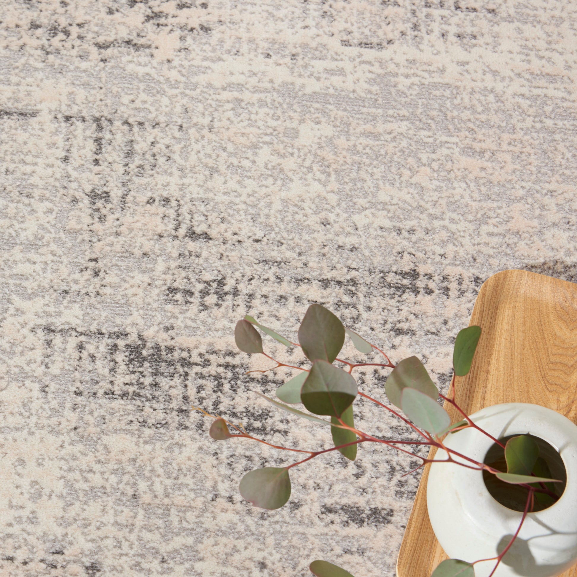 Beige and gray abstract rug with a light wood tray and white ceramic vase holding eucalyptus stems with red-tinted leaves.