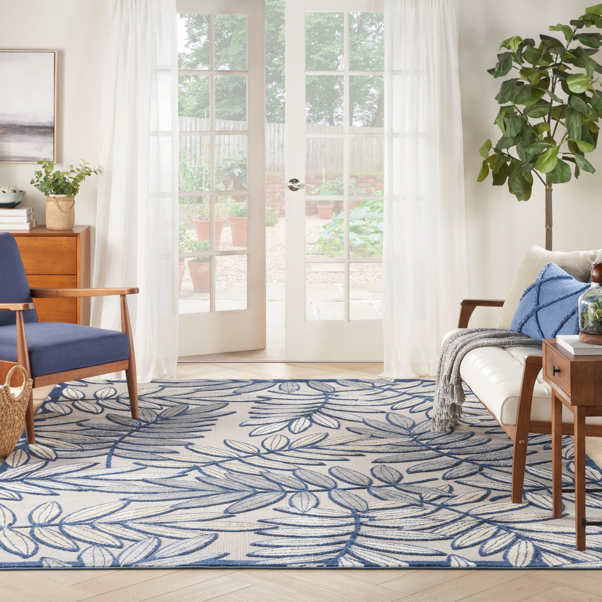 Bright living room with white sheer curtains, blue leaf rug, wooden coffee table, sofa with cushions, and a potted plant.
