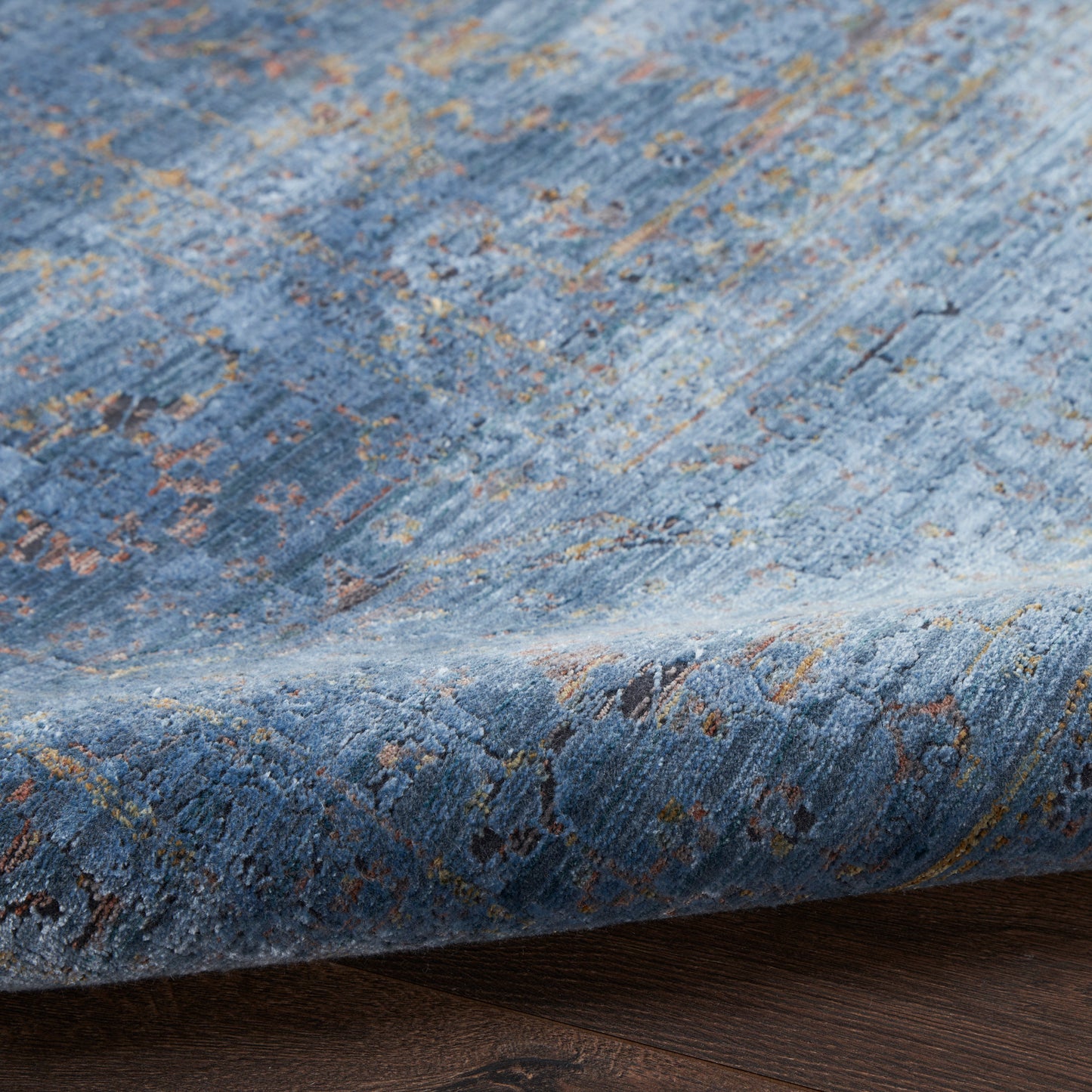 Blue distressed oriental rug with gold & brown floral motifs, plush pile, partially folded over dark wood floor.
