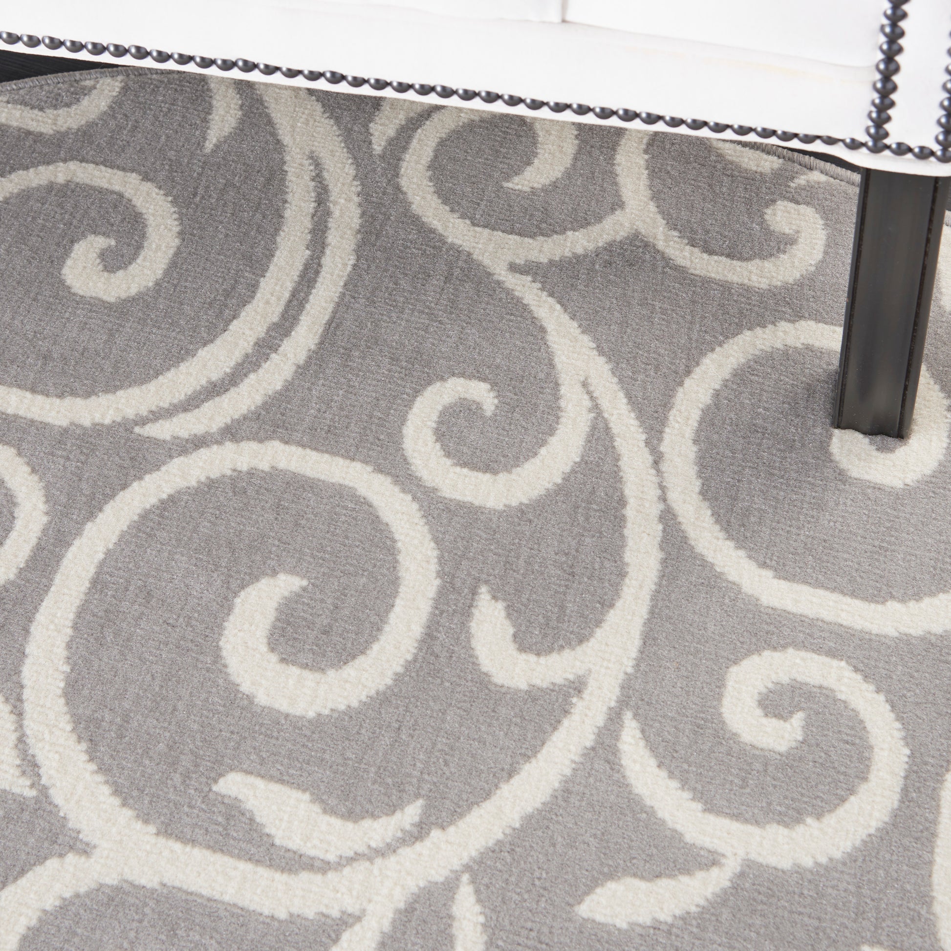 Gray rug with cream scrolling damask pattern; white upholstered chair edge with nailhead trim and black leg.