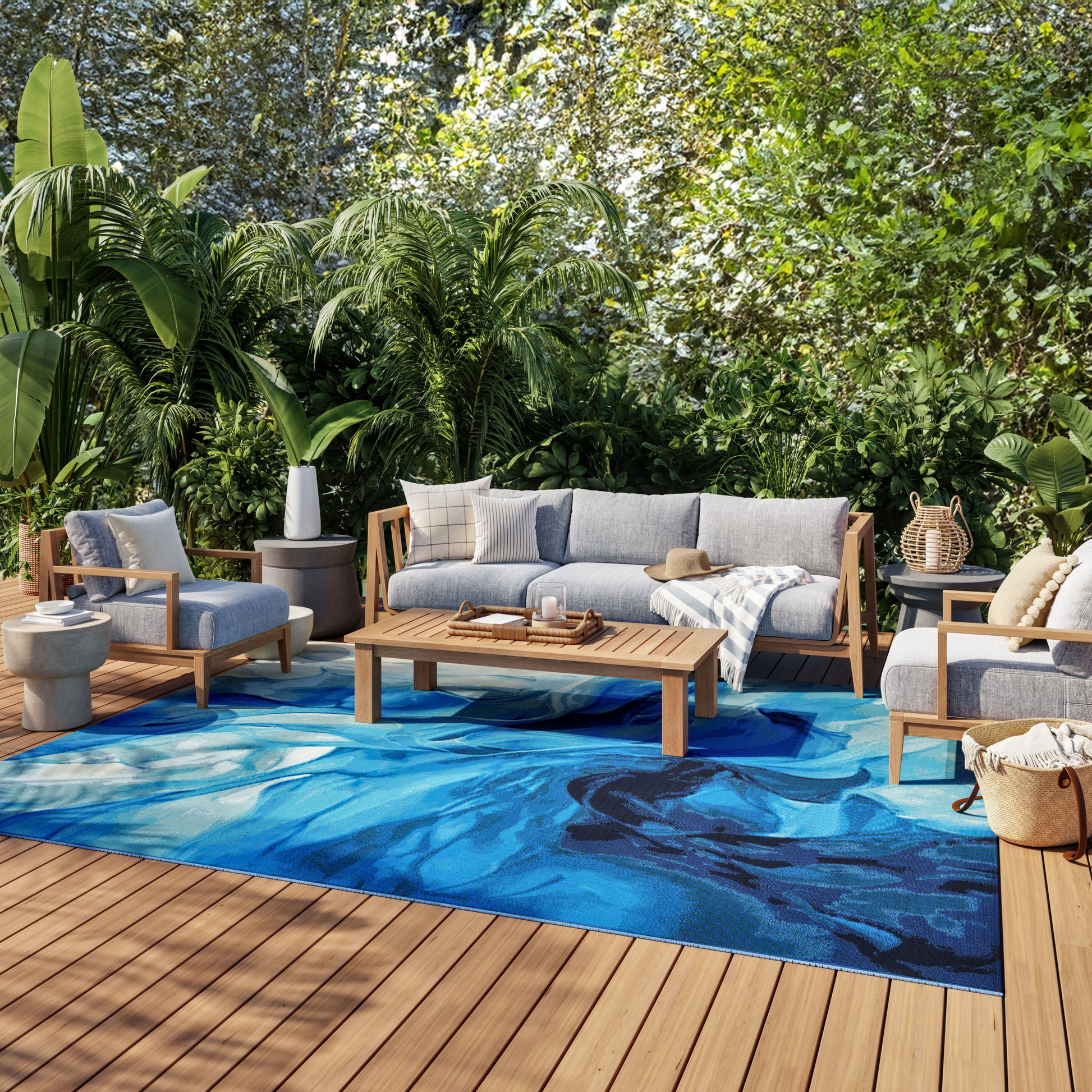 Outdoor lounge on a wood deck with gray cushioned sofa and chairs, blue abstract rug, coffee table, tropical plants.