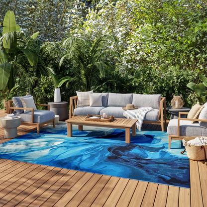 Outdoor lounge on a wood deck with gray cushioned sofa and chairs, blue abstract rug, coffee table, tropical plants.