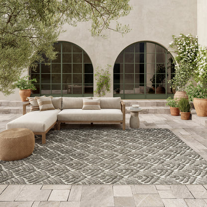 Outdoor lounge with beige fabric sectional, light stone patio, grey patterned outdoor rug, woven pouf, potted plants, and green arched window backdrop.