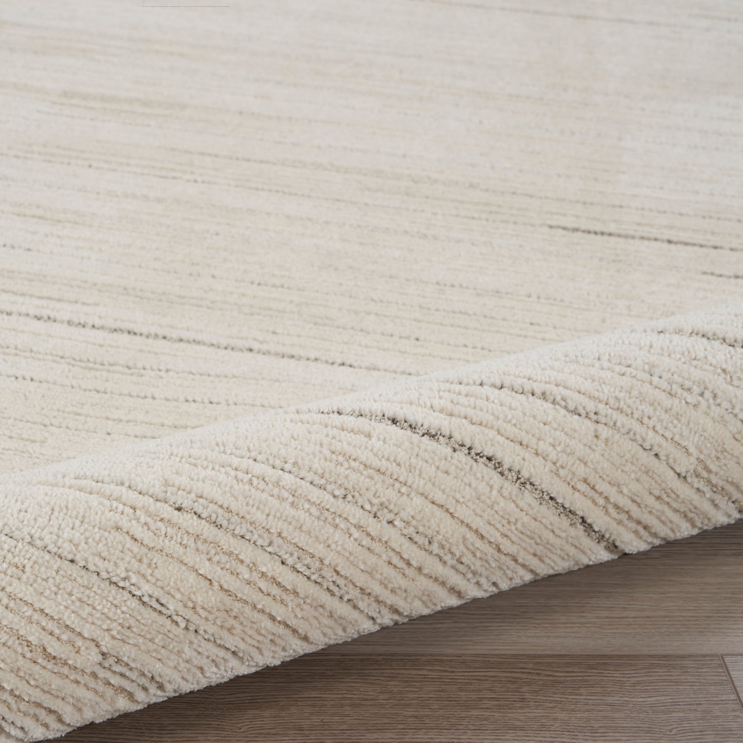 Cream beige shag rug with subtle linear stripes and plush pile, curled edge on light wood flooring.