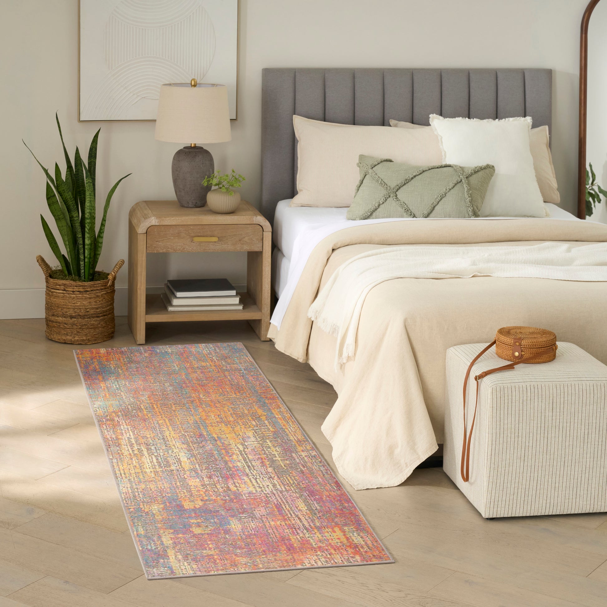 Cozy bedroom with gray upholstered bed, beige bedding, multicolor runner rug, wood nightstand, lamp and potted plant.