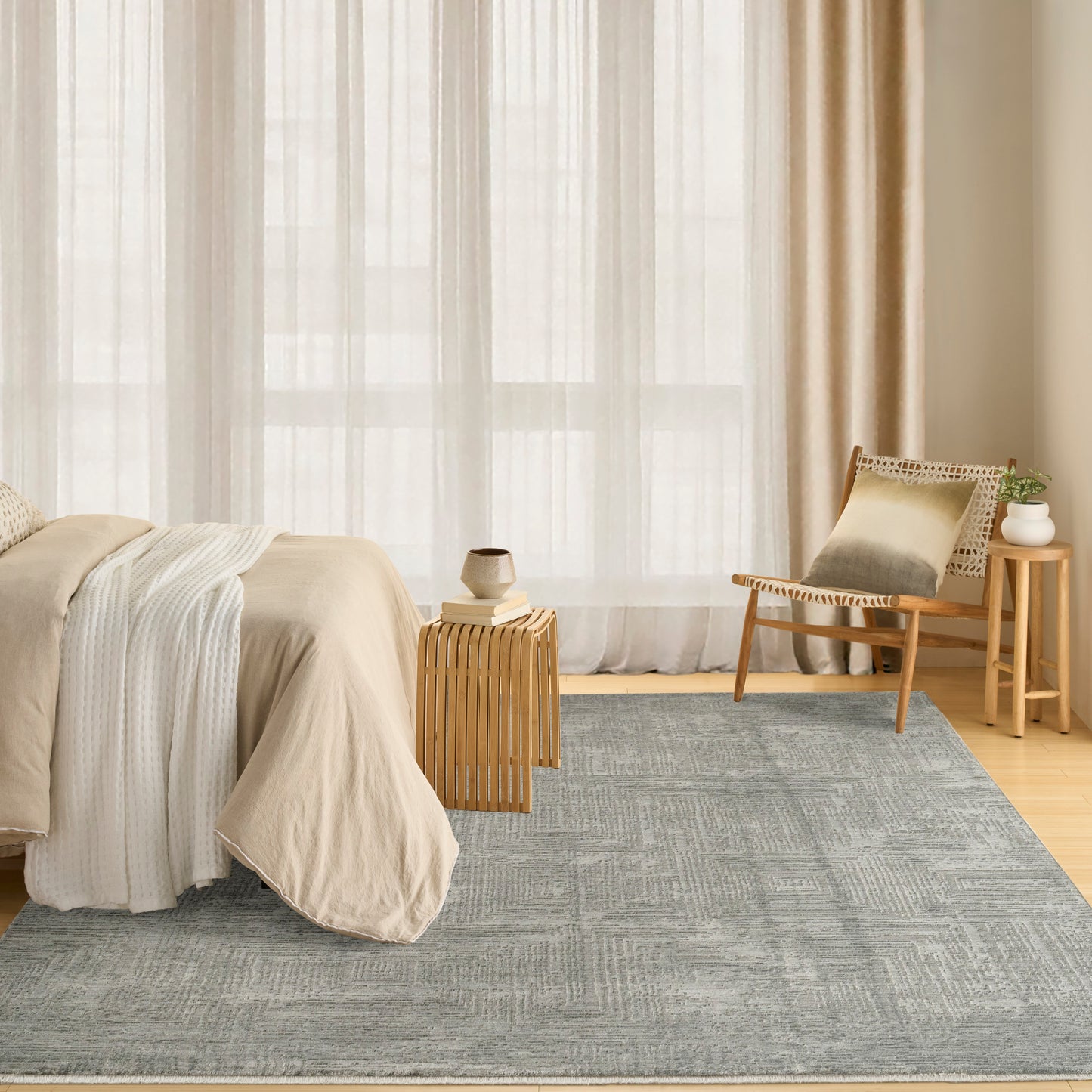 Neutral-toned bedroom featuring a light gray-blue rug, beige duvet, white throw, sheer beige curtains, wooden chair and a small plant on a side table.