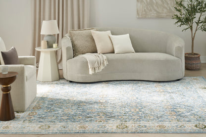 Curved light gray-beige fabric sofa with beige cushions and throw, in a neutral living room with a blue-toned patterned rug.