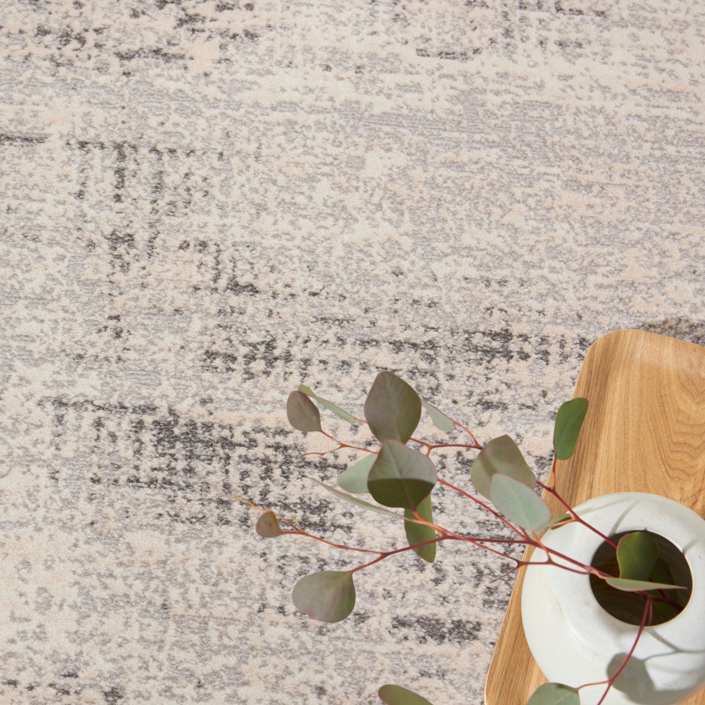 Beige and gray patterned rug with a light wood tray and white ceramic vase holding eucalyptus stems and round green leaves.