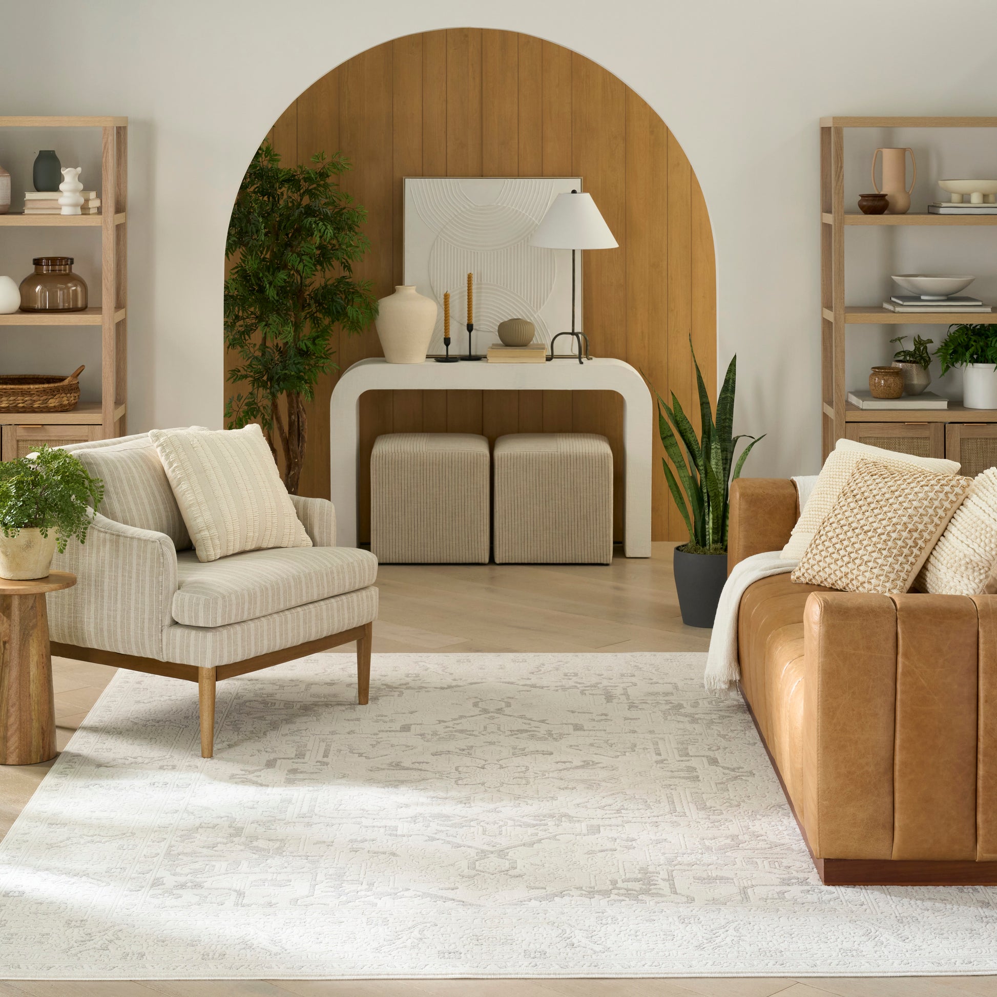 Neutral rug in a living room with an arched wooden backdrop, white console, beige chairs, brown leather sofa, potted plants and open shelves.