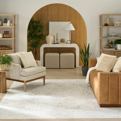 Neutral rug in a living room with an arched wooden backdrop, white console, beige chairs, brown leather sofa, potted plants and open shelves.