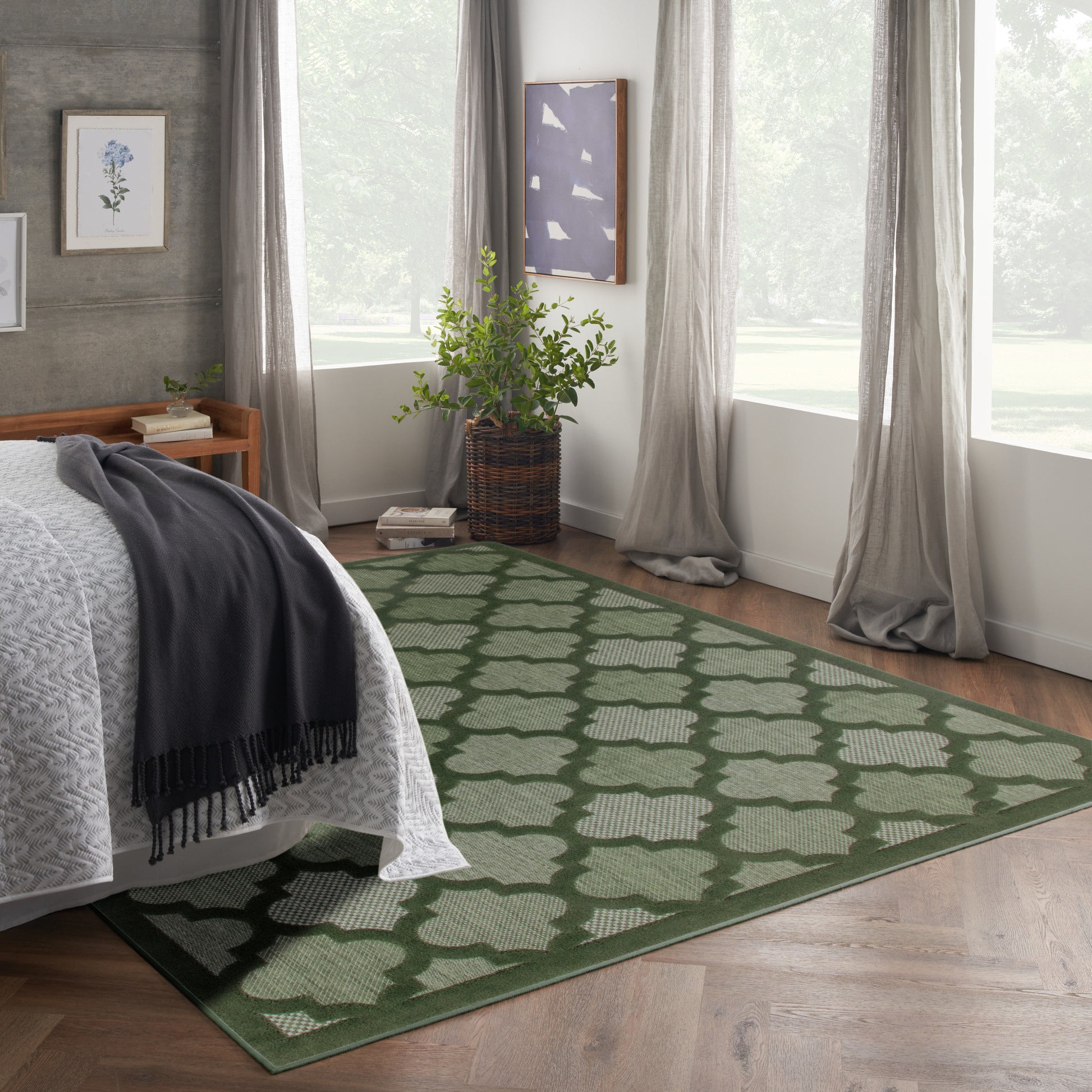Bedroom with green geometric rug on wood floor, bed with white coverlet and dark gray throw, plant in wicker basket.