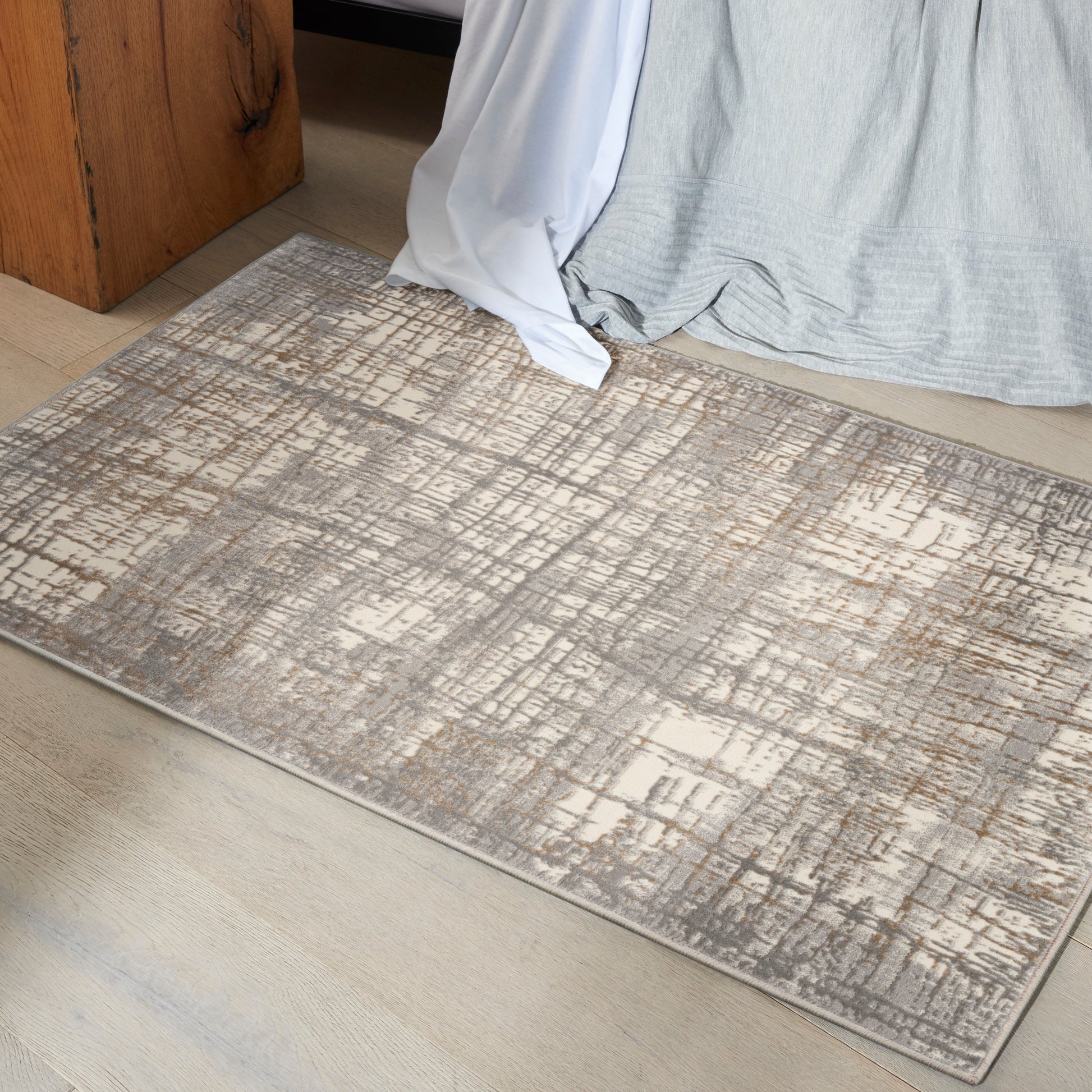 Neutral abstract-pattern area rug in gray and beige on light wood floor beside bed with draped gray and white linens.