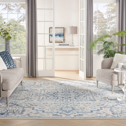Light gray fabric sofas frame a blue-and-white rug in a bright living room with glass doors, plants, and gray curtains.