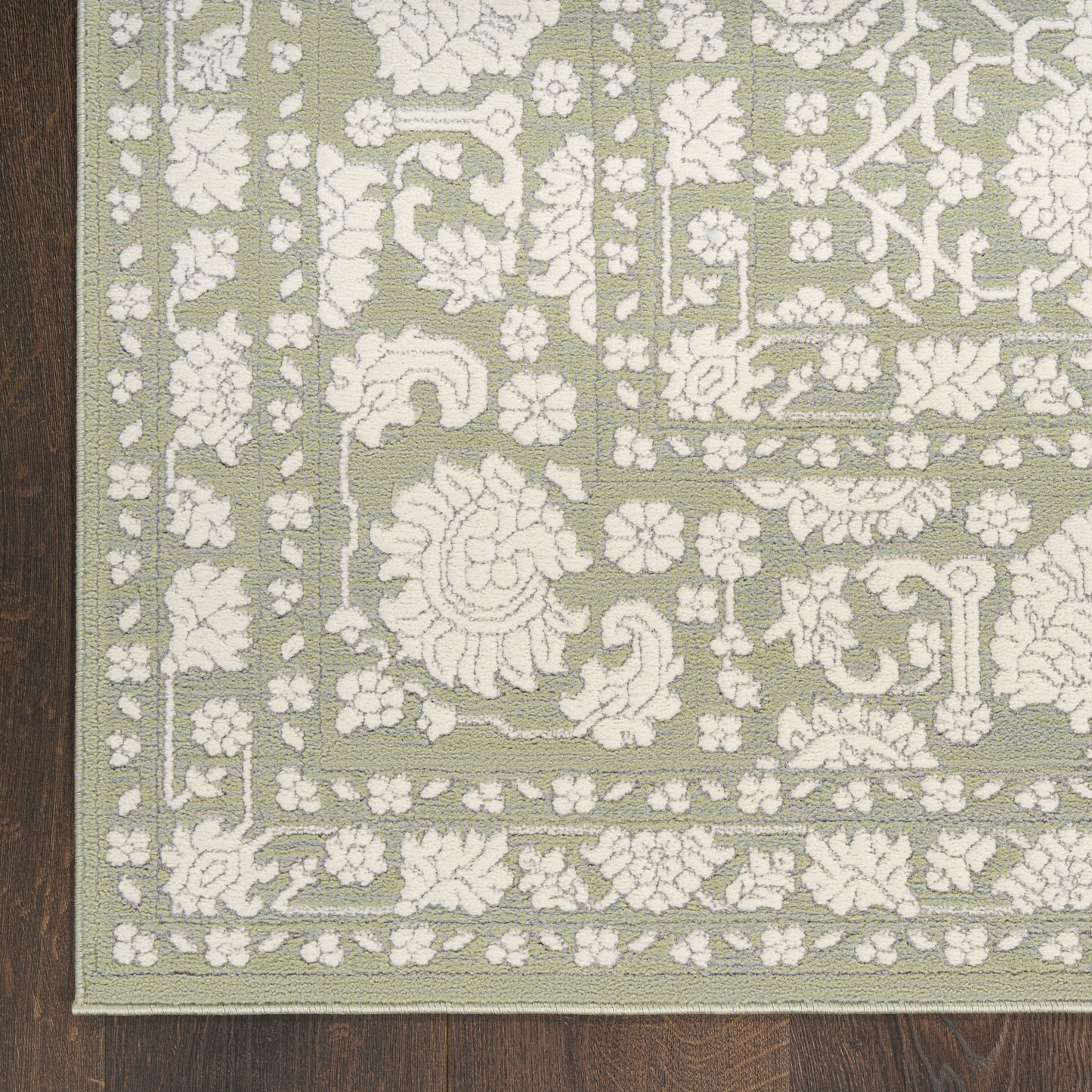 Rectangular sage green rug with white raised floral pattern and decorative border on dark wood floor.