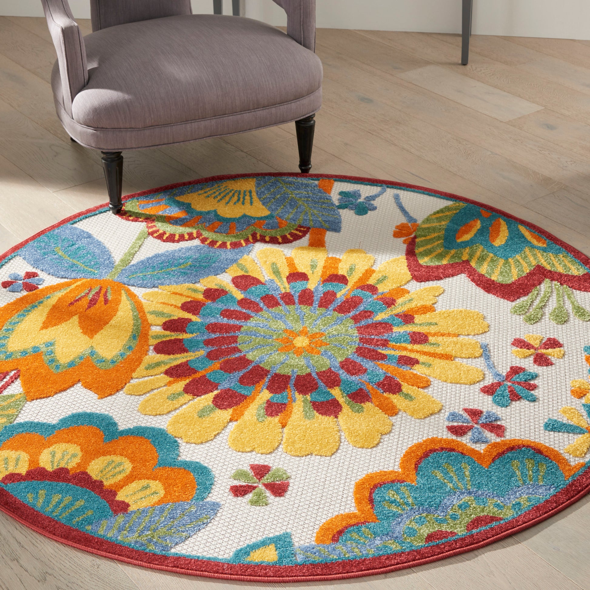 Round multicolor floral rug with yellow, blue and orange petals, red border on a wood floor beside a purple chair.