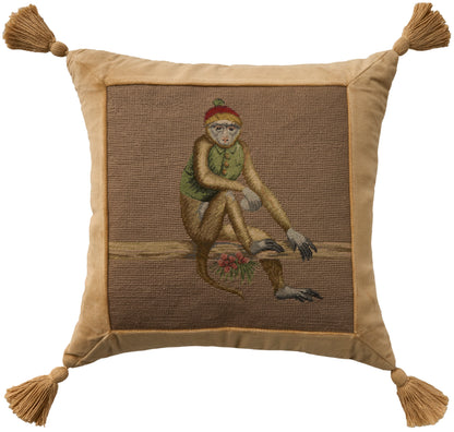 Gold-tan square pillow with tassel corners; embroidered lounging figure on brown panel, wearing green top and red hat.
