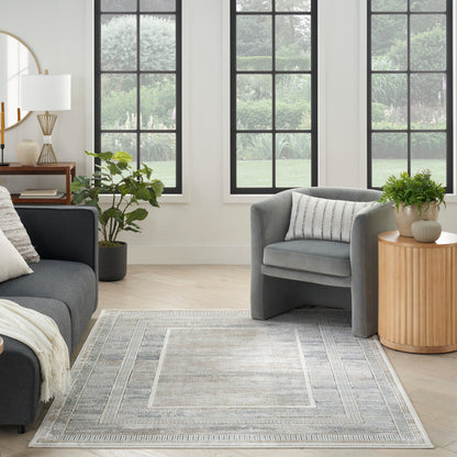 Modern living room with grey sofas, a patterned light gray rug, round wooden side table, green plants, and large windows.