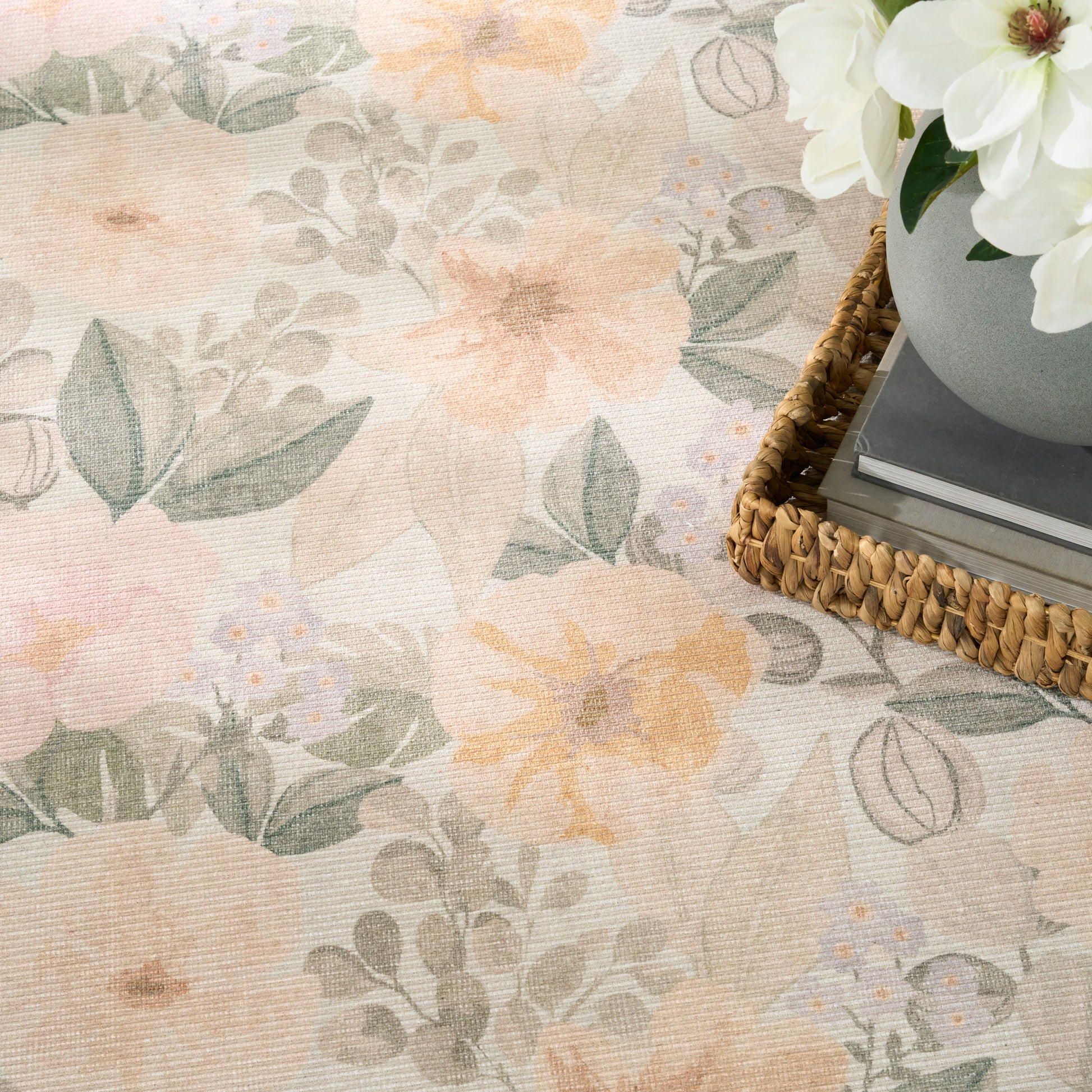 Pastel floral rug with peach blossoms and gray-green leaves; woven wicker tray with books and a vase of white flowers.