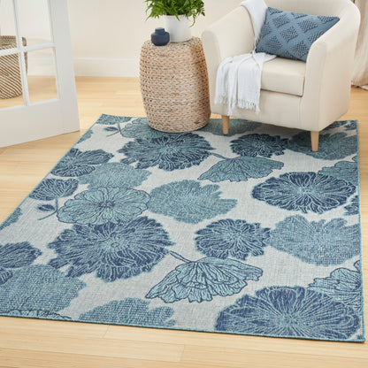 Blue floral area rug on light wood floor beside cream upholstered armchair and woven rattan side table with plant.