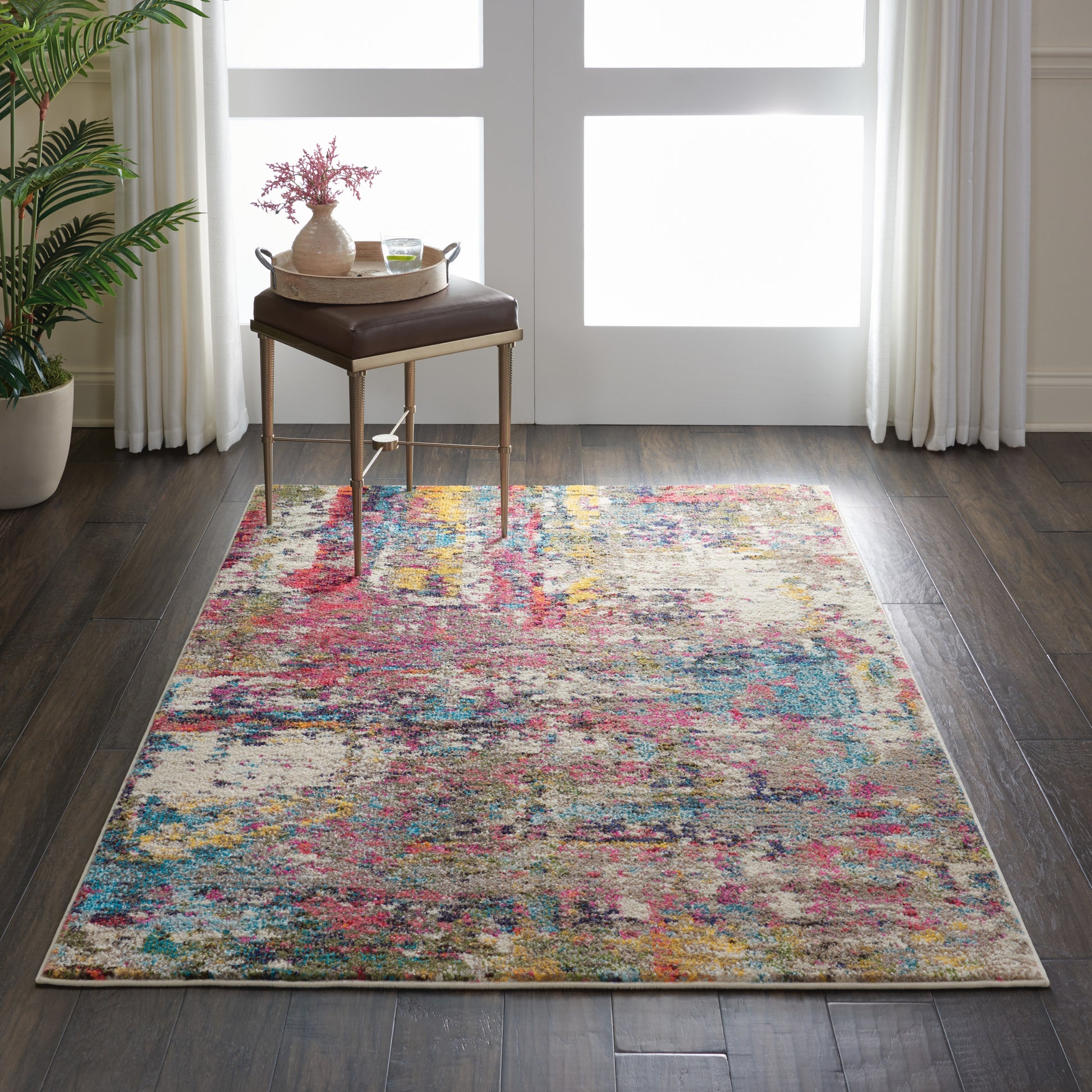 Multicolor abstract area rug on dark wood floor, brown cushioned stool and round wooden tray by window.
