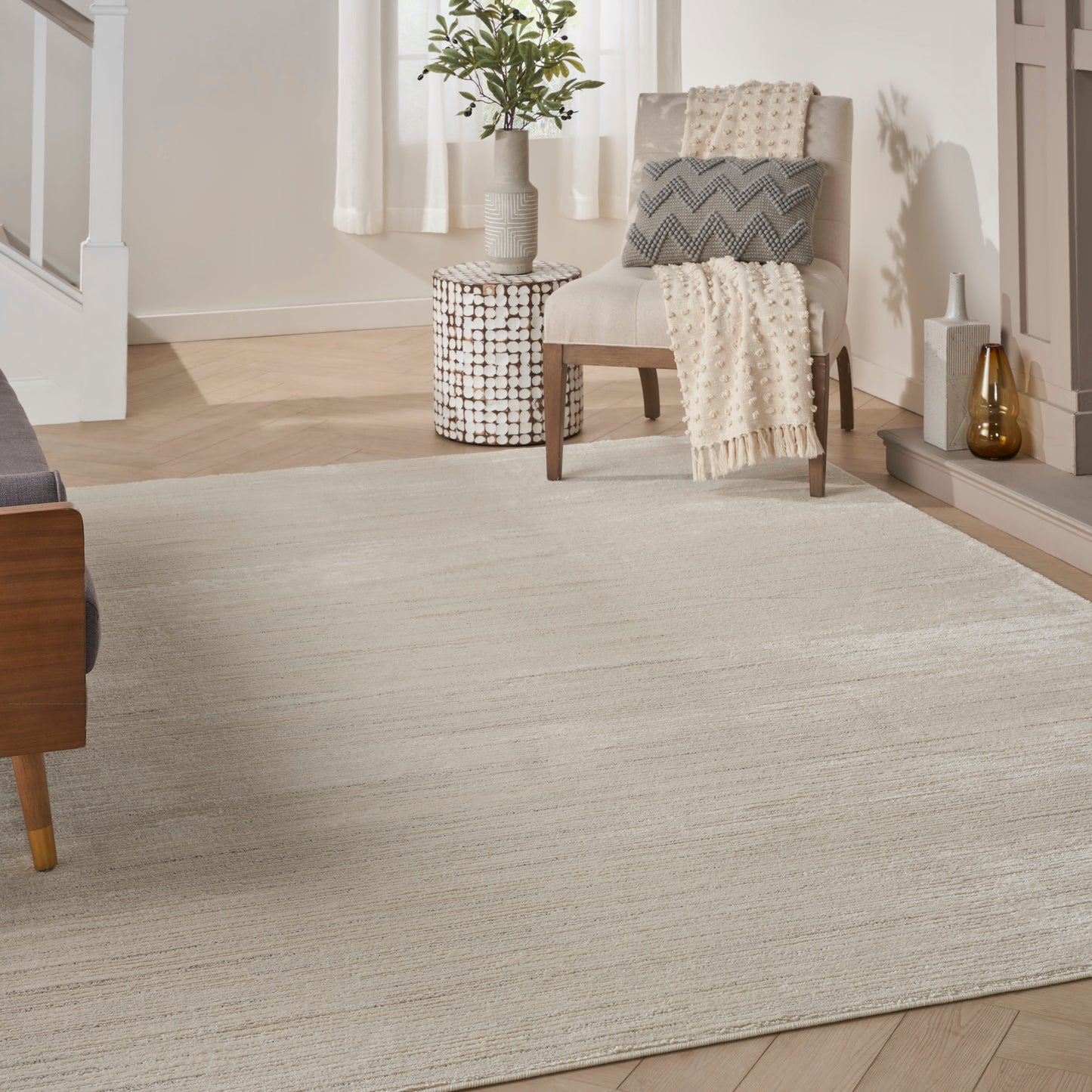 Beige woven area rug on light wood floor; cream chair with knit throw, potted plant on woven stool.