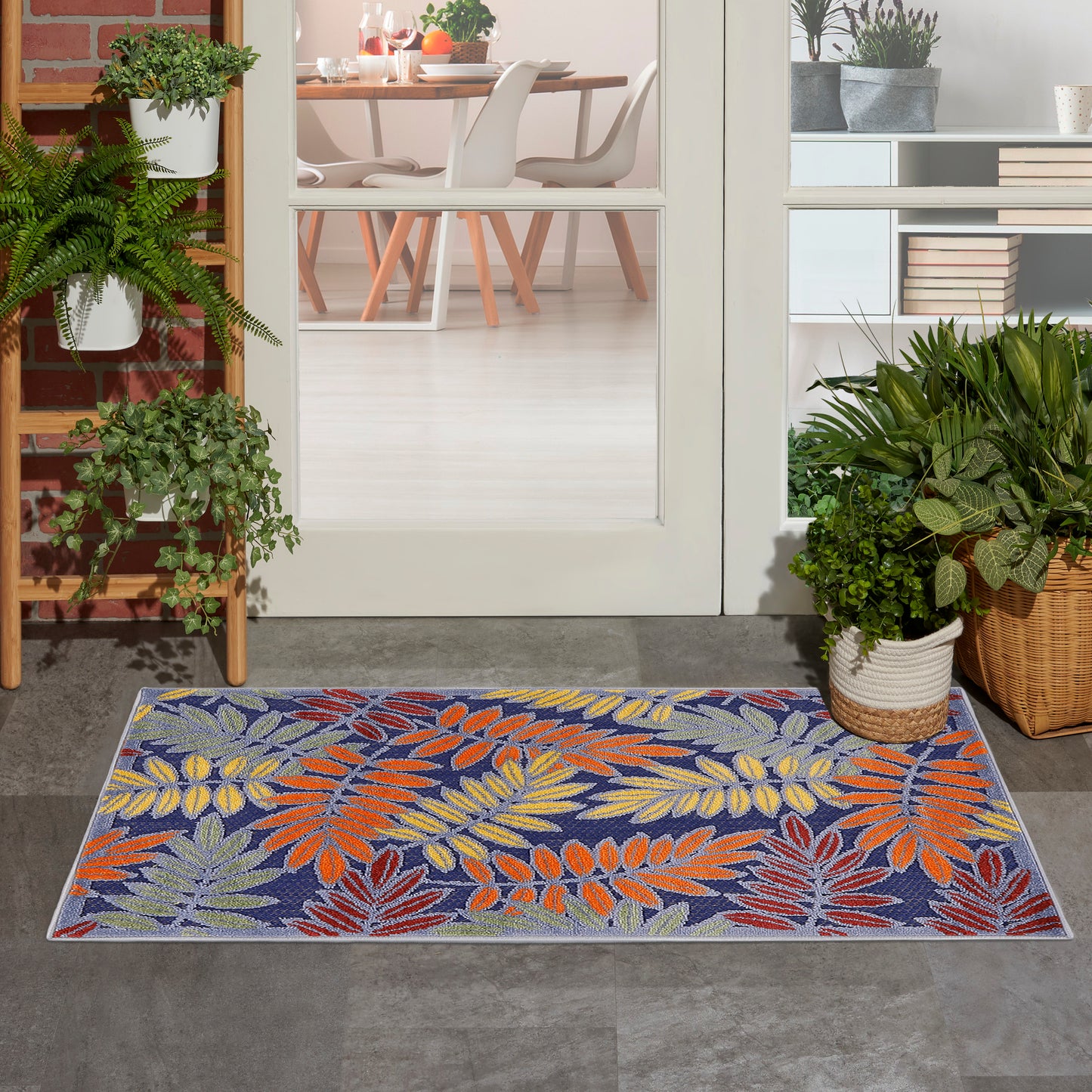 Outdoor rug with a tropical leaf pattern in orange, yellow, and purple on a blue background at a doorway with potted plants.