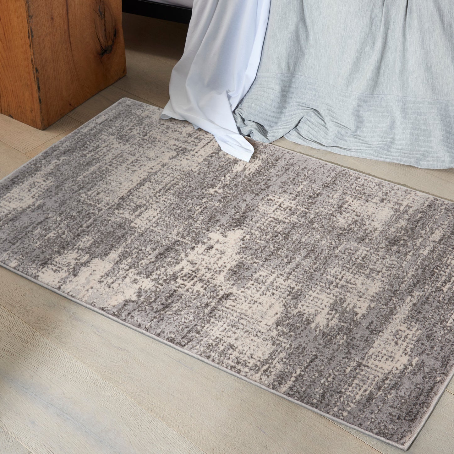 Rectangular gray-beige area rug with an abstract pattern on a light wood floor beside a bed.