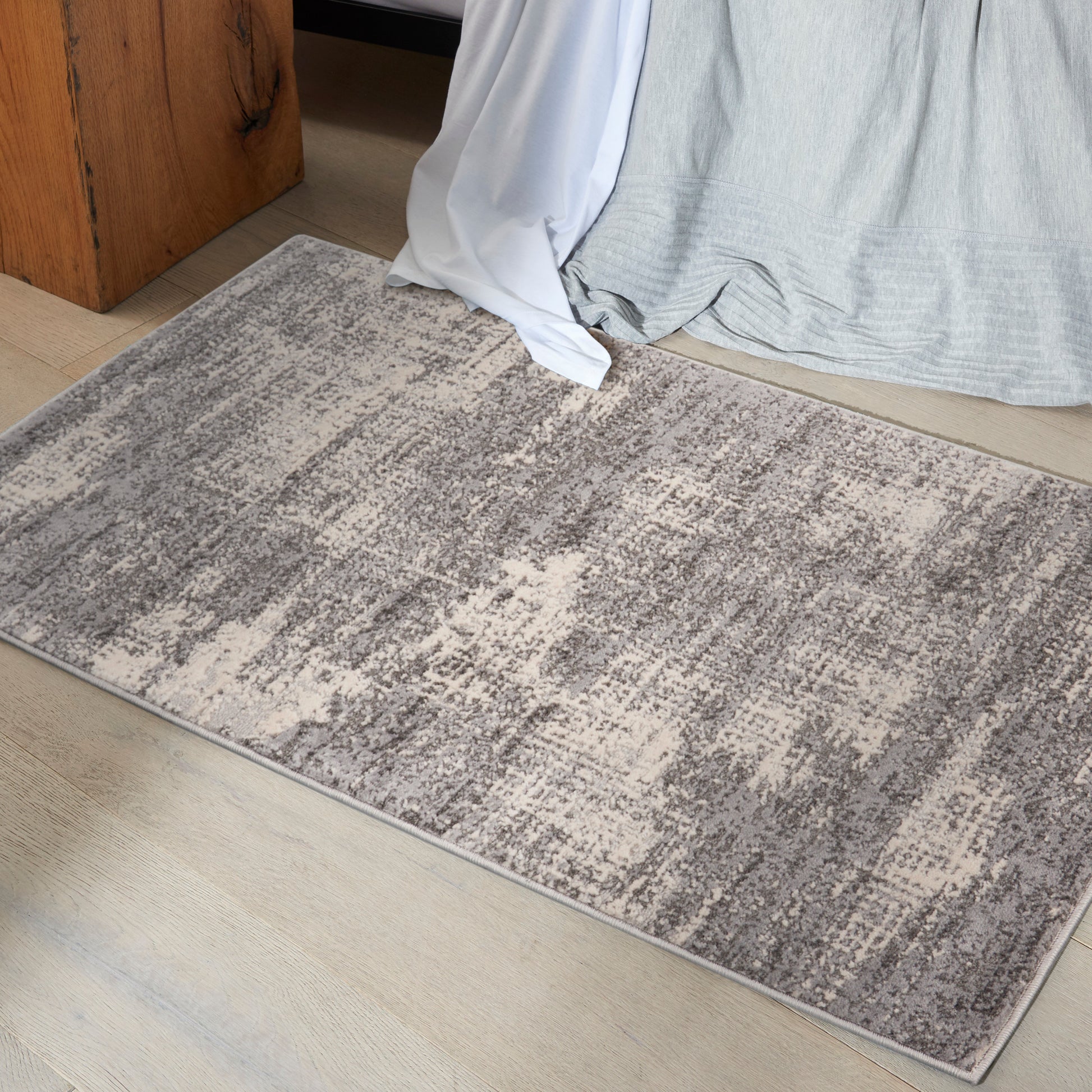 Rectangular gray-beige area rug with an abstract pattern on a light wood floor beside a bed.