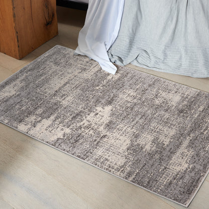Rectangular gray-beige area rug with an abstract pattern on a light wood floor beside a bed.