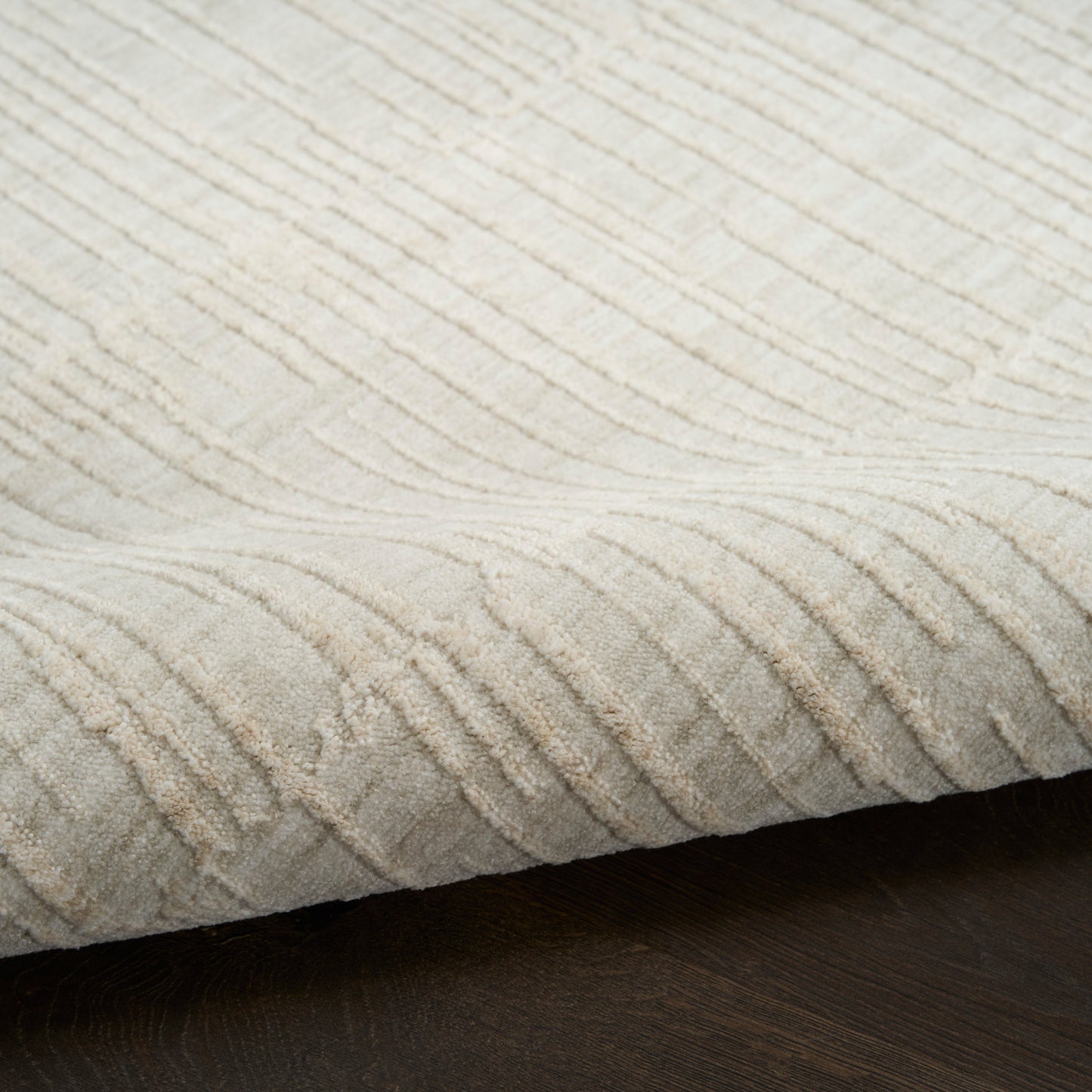 Beige textured rug with soft ribbed, wavy pattern; edge folded over dark wooden floor.