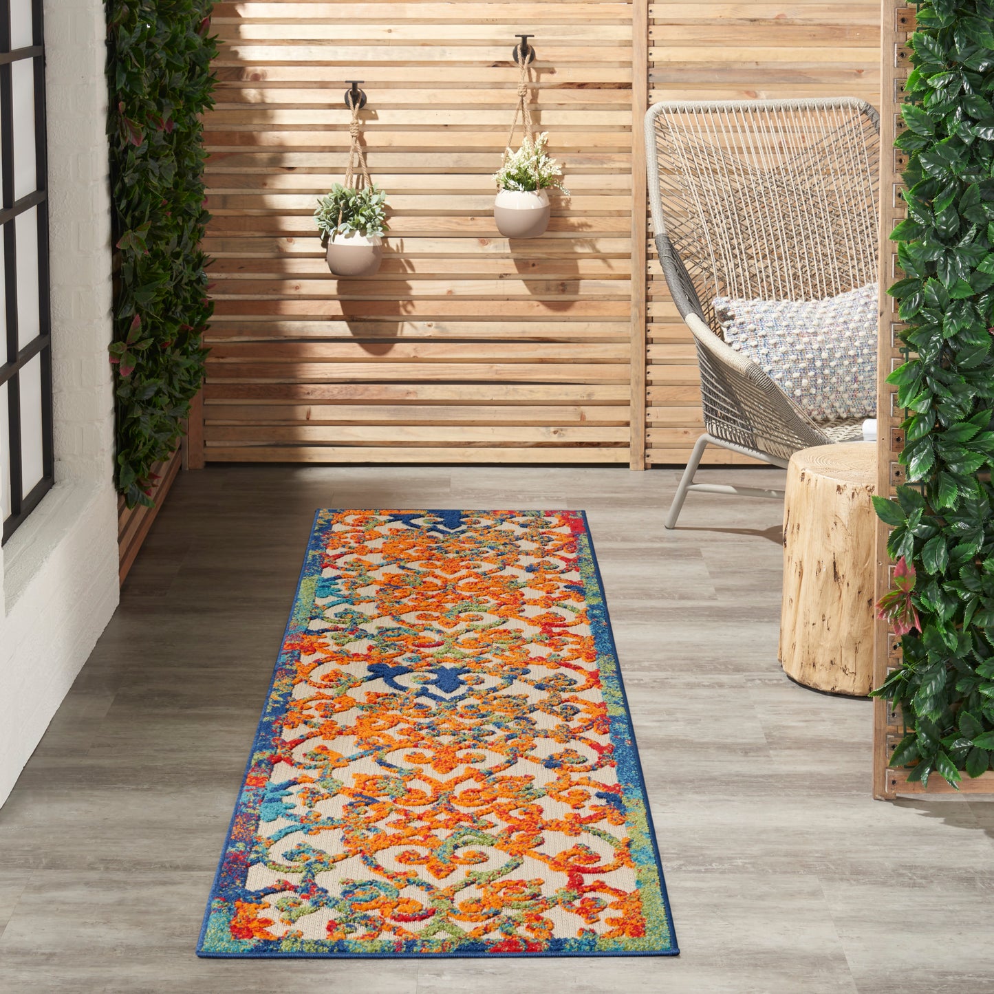 Outdoor runner rug with orange florals and blue border on a wood deck beside a wicker chair and hanging planters.