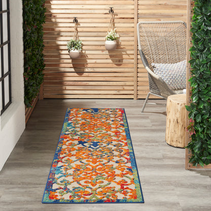 Outdoor runner rug with orange florals and blue border on a wood deck beside a wicker chair and hanging planters.
