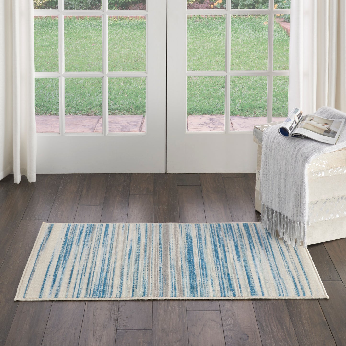 Rectangular blue striped area rug on dark wood floor before glass French doors; cream sofa with throw and magazine.