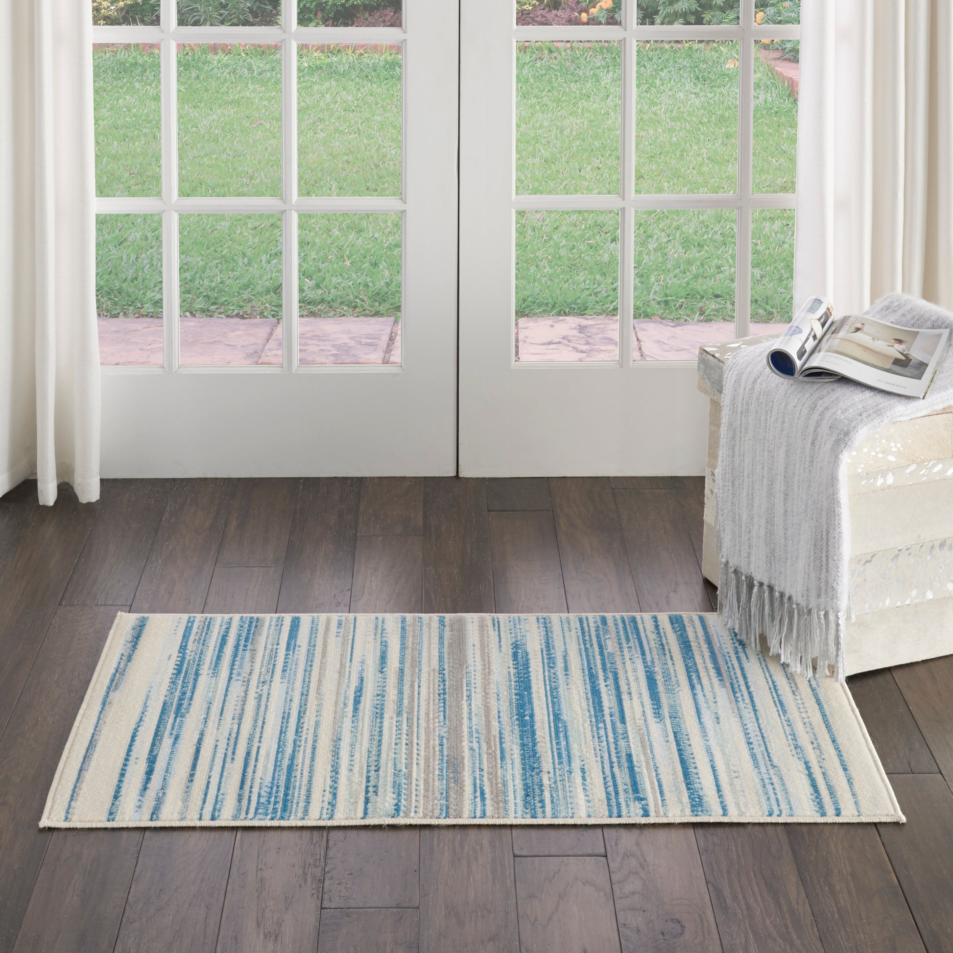 Rectangular blue striped area rug on dark wood floor before glass French doors; cream sofa with throw and magazine.
