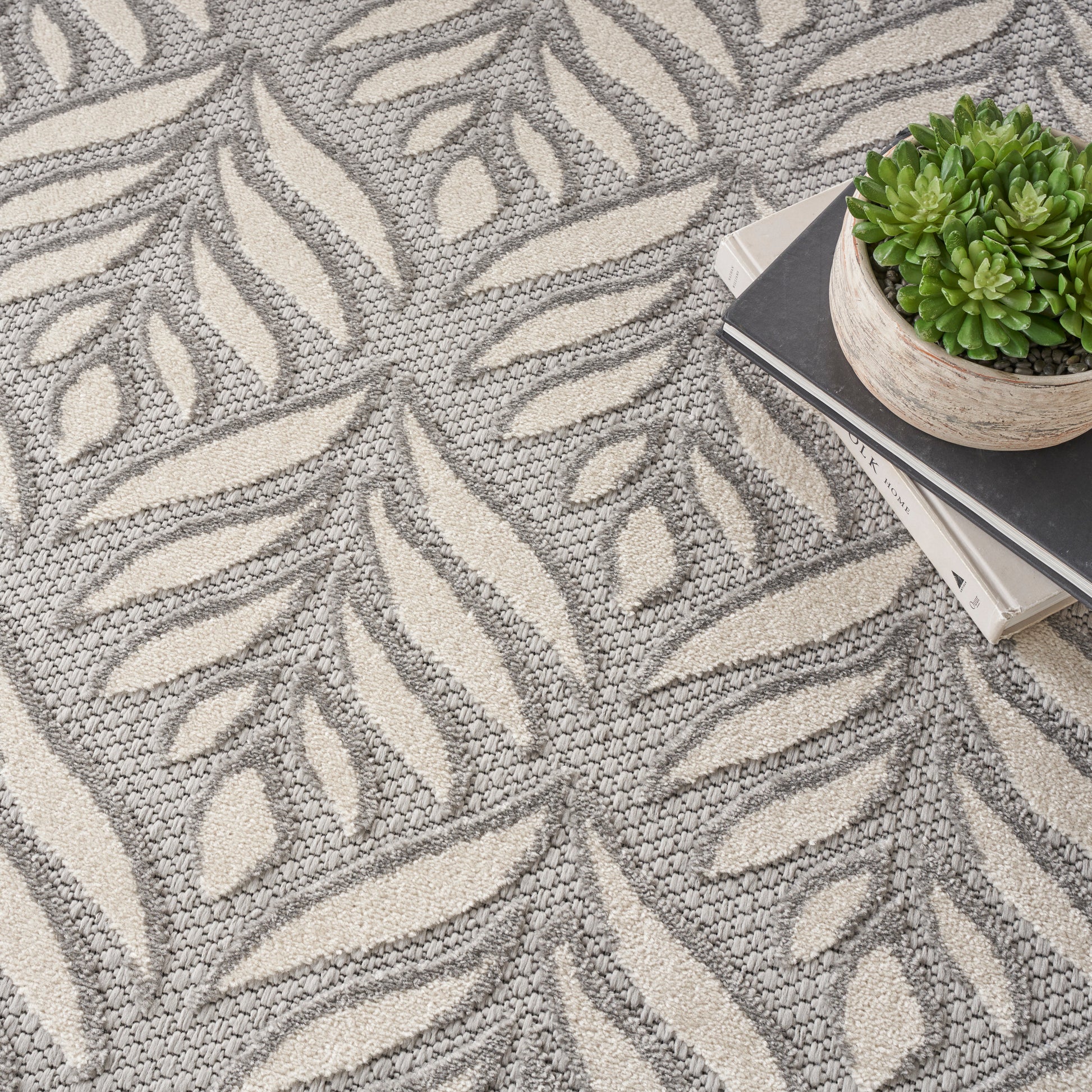 Gray and cream leaf-patterned woven rug; modern botanical design with a stack of books and a small potted succulent.
