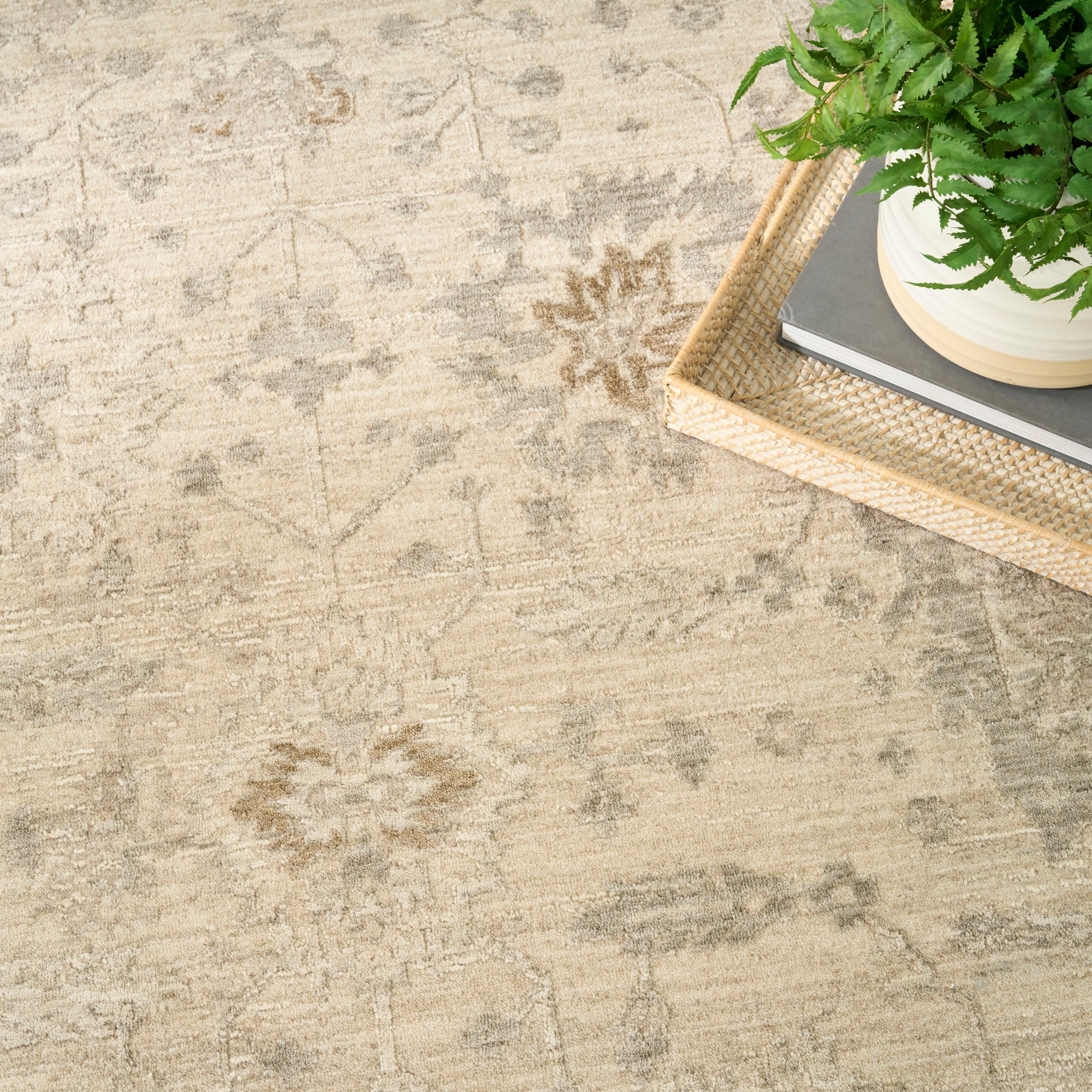 Beige oriental rug with subtle floral motif; light wicker tray holds a potted green fern and stacked books.