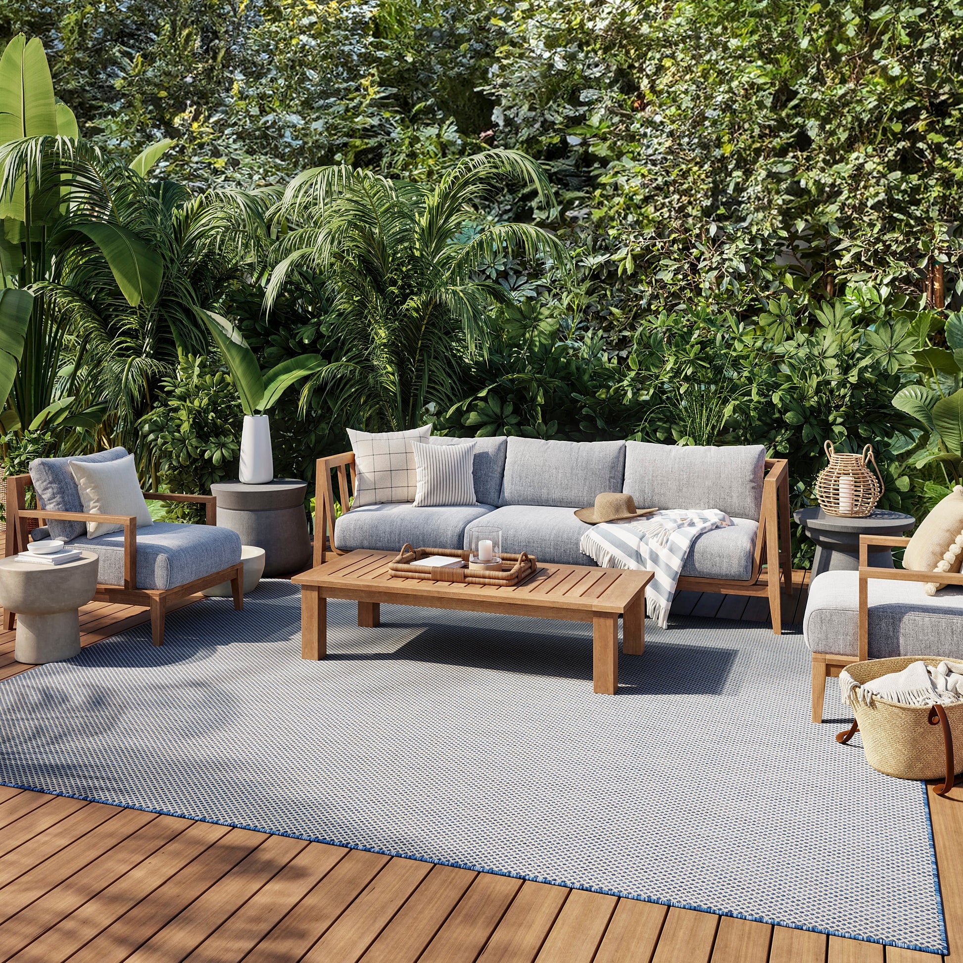 Outdoor lounge set with gray cushions and wooden frames, coffee table on a blue-gray rug amid tropical plants.